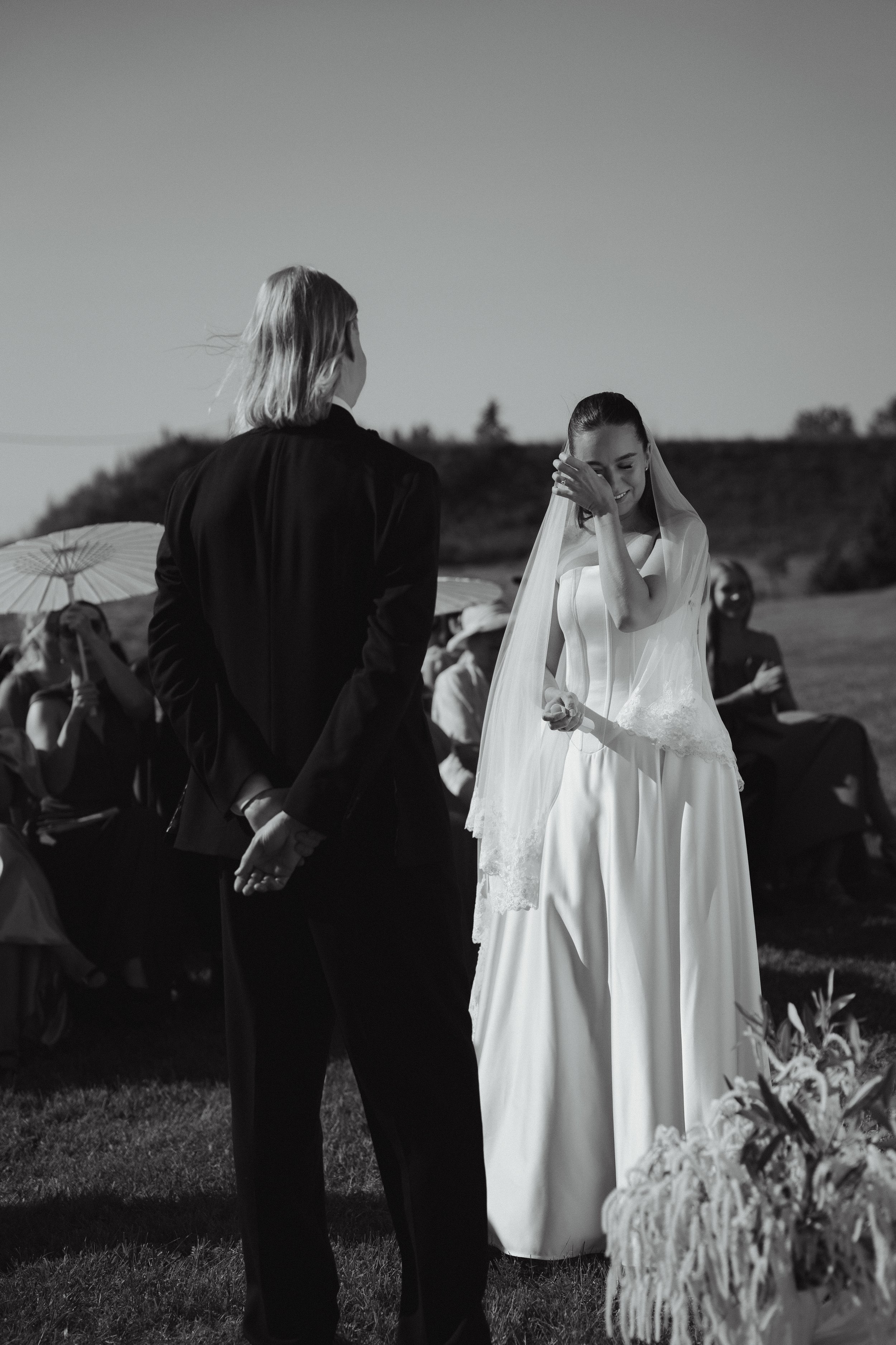 A black and white photo of a wedding ceremony outdoors, with a bride in a wedding dress and veil holding a flower, and a groom in a suit standing in front of her. Guests are seated in the background.