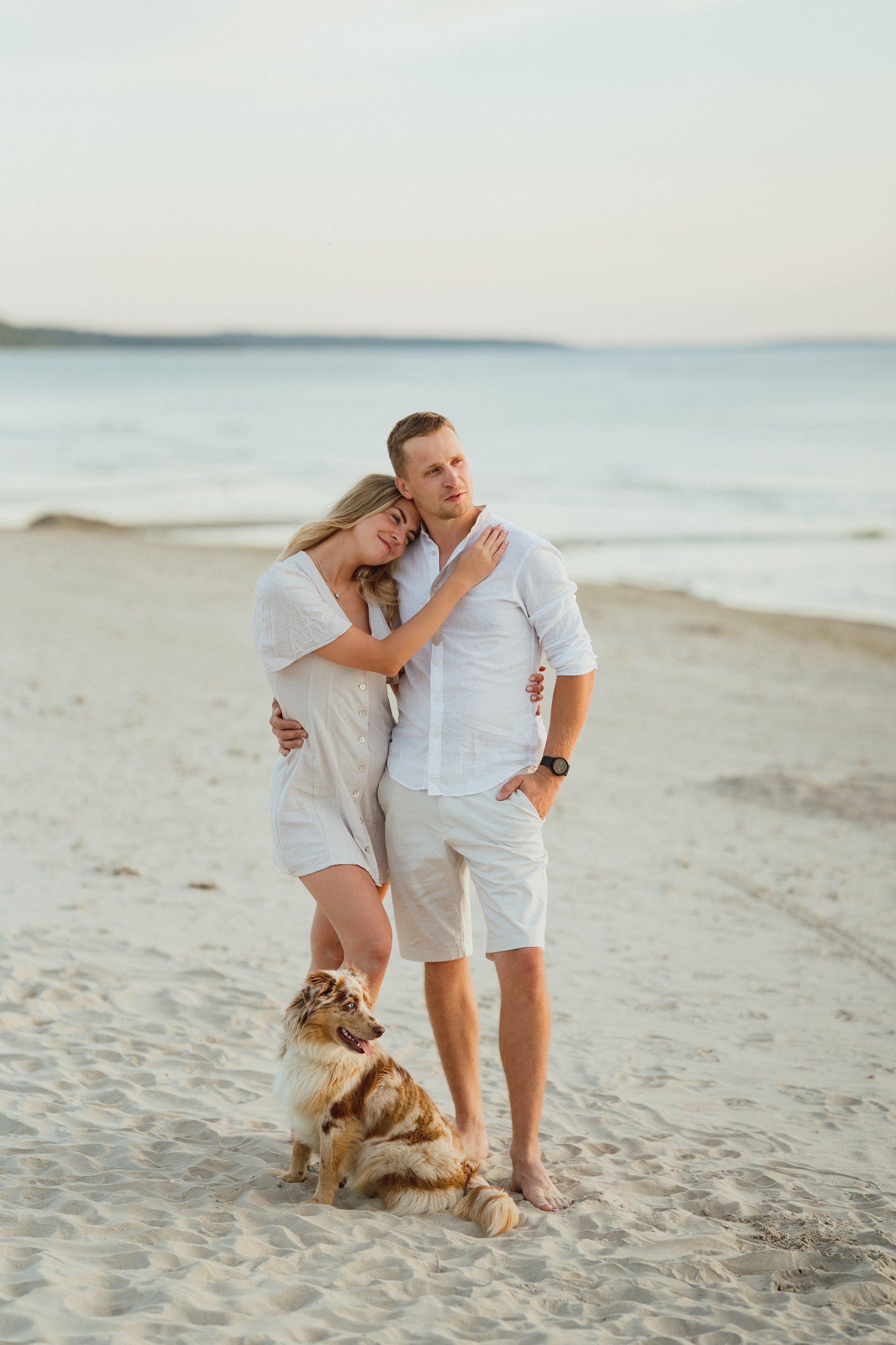A couple and a dog on a sandy beach with the ocean in the background, during sunset.