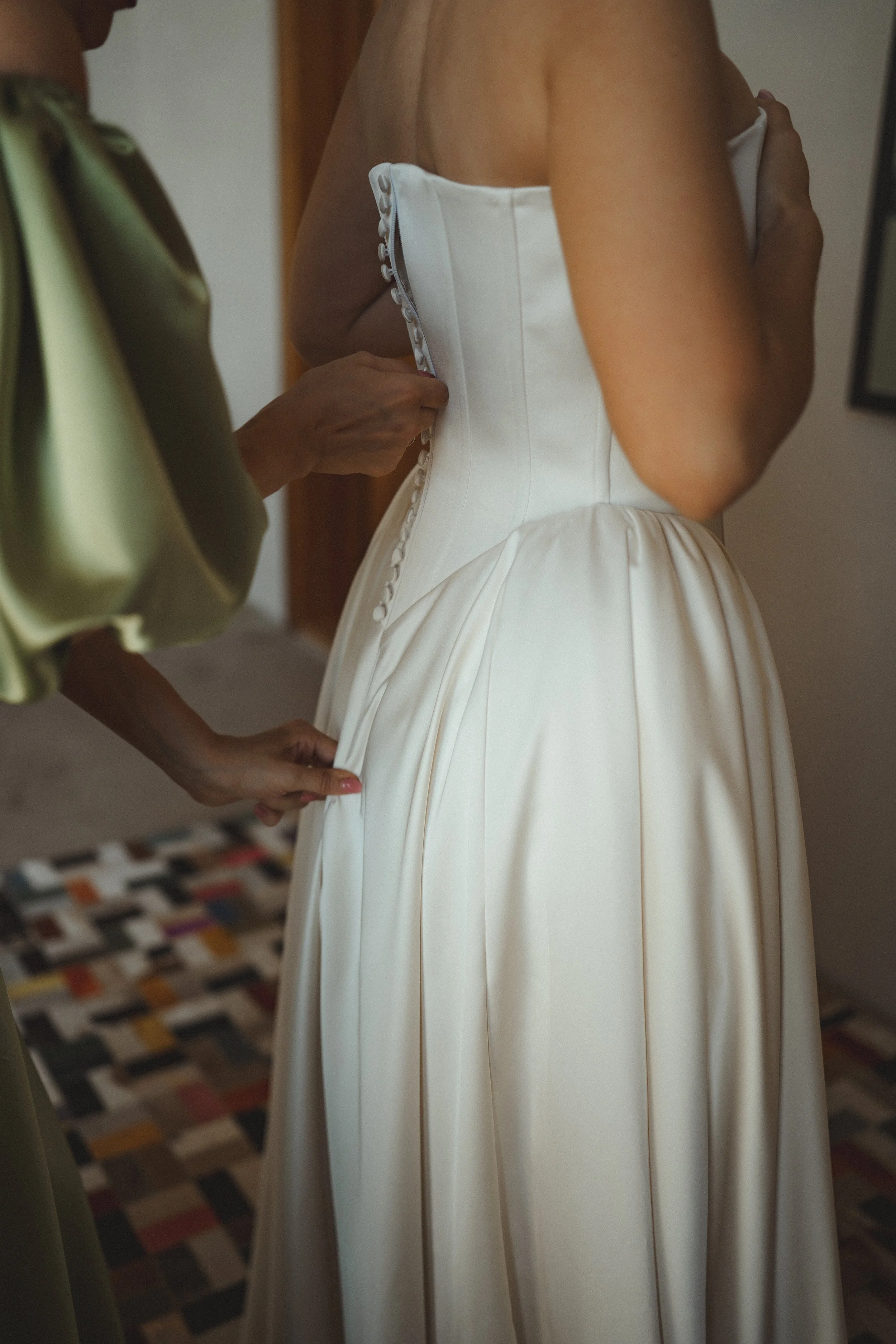 A bride in a white strapless wedding dress with a full skirt is being assisted in buttoning her dress.