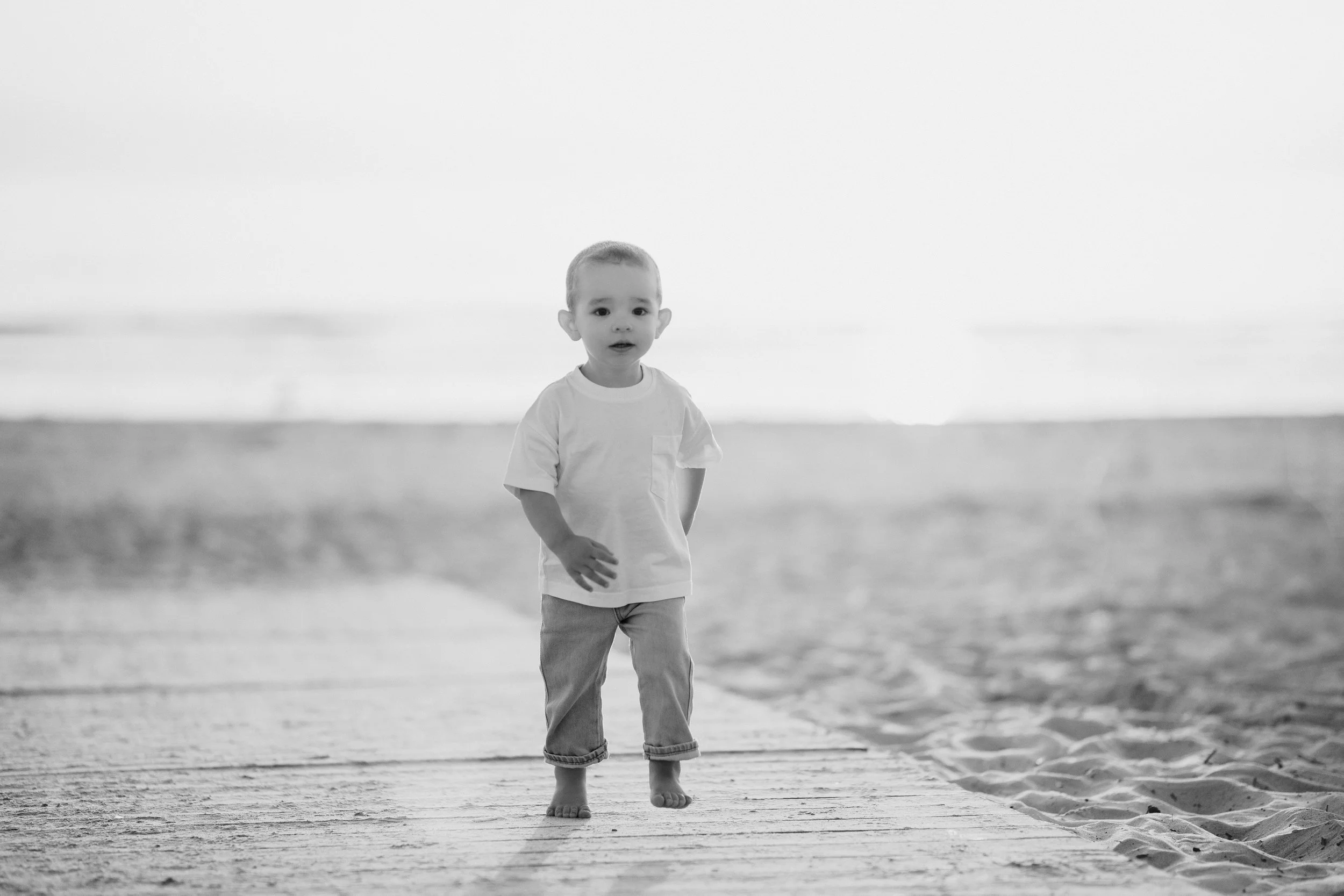 A young boy standing barefoot on a wooden pier at the beach, looking at the camera with a surprised expression, in black and white.