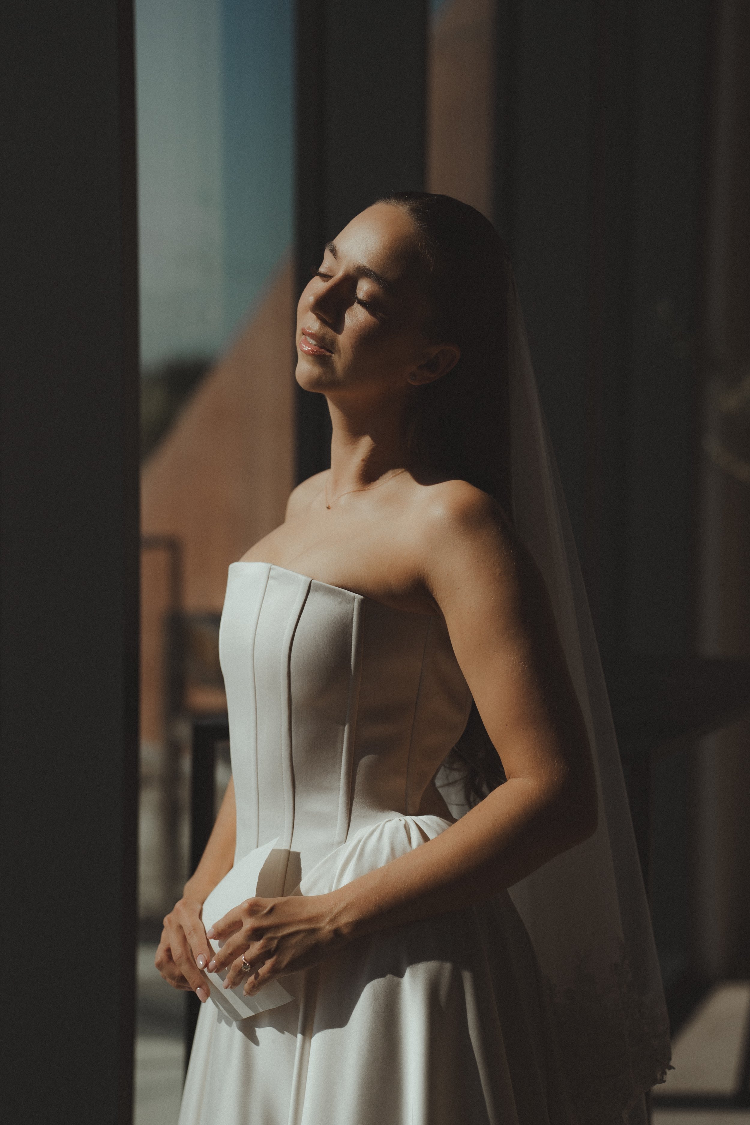 Bride in a strapless white wedding dress standing with eyes closed, facing sunlight inside a building.