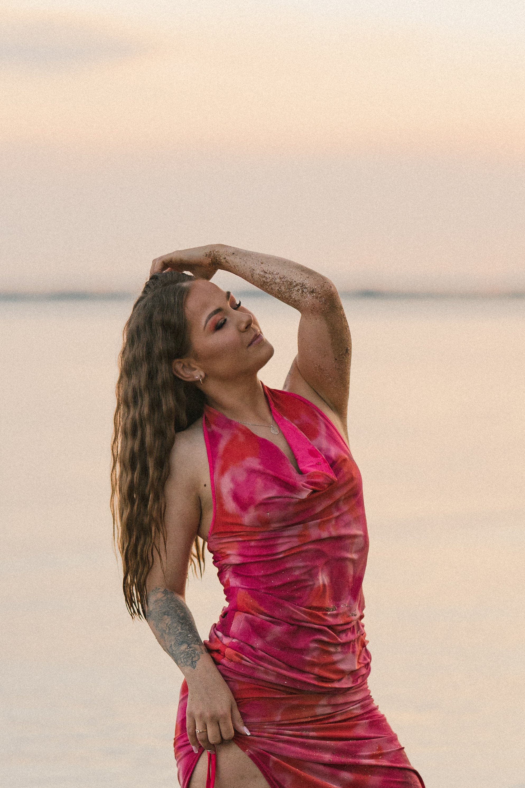 A woman with long wavy hair and a colorful dress with hints of pink and orange stands with her eyes closed and her hand on her head in front of a calm body of water at sunset.