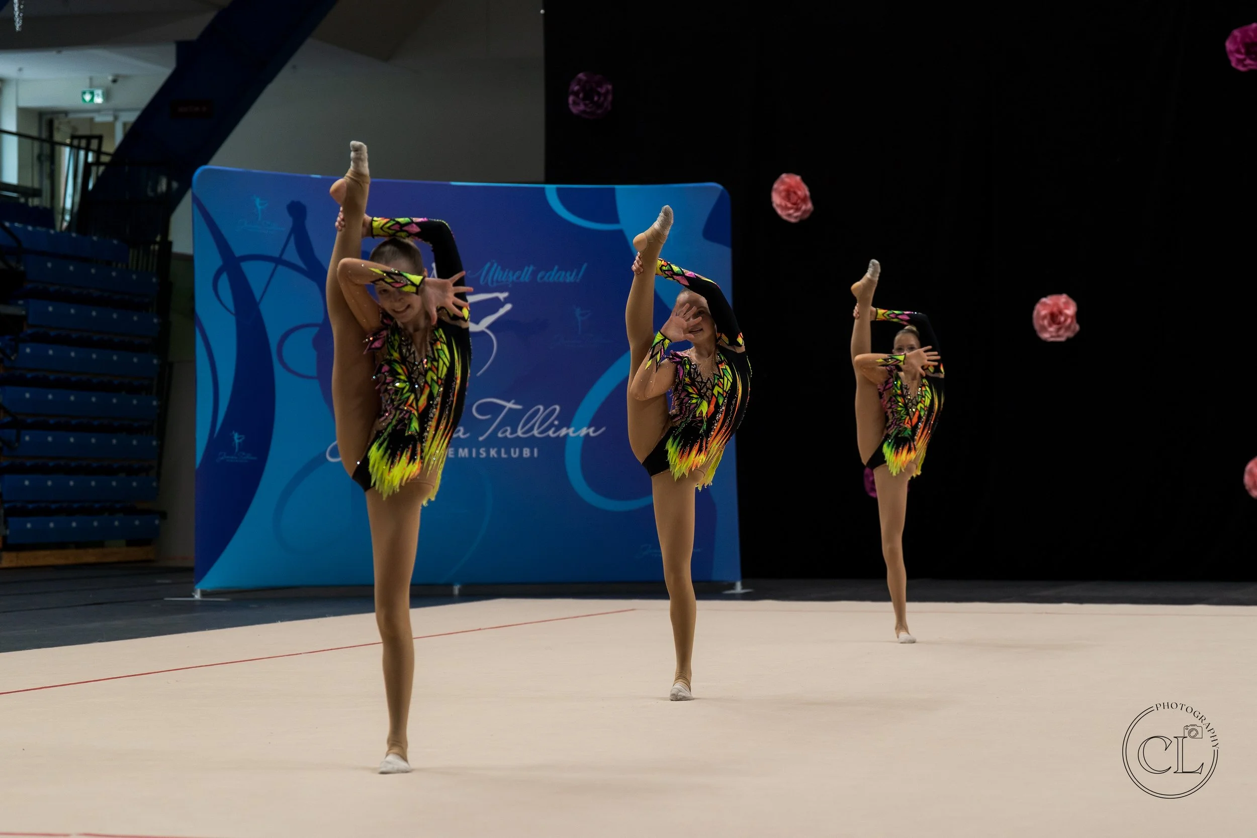 Three young female gymnasts in colorful black, yellow, and pink leotards performing a synchronized routine on a beige carpeted floor during a competition, with a blue backdrop and pink flower decorations.