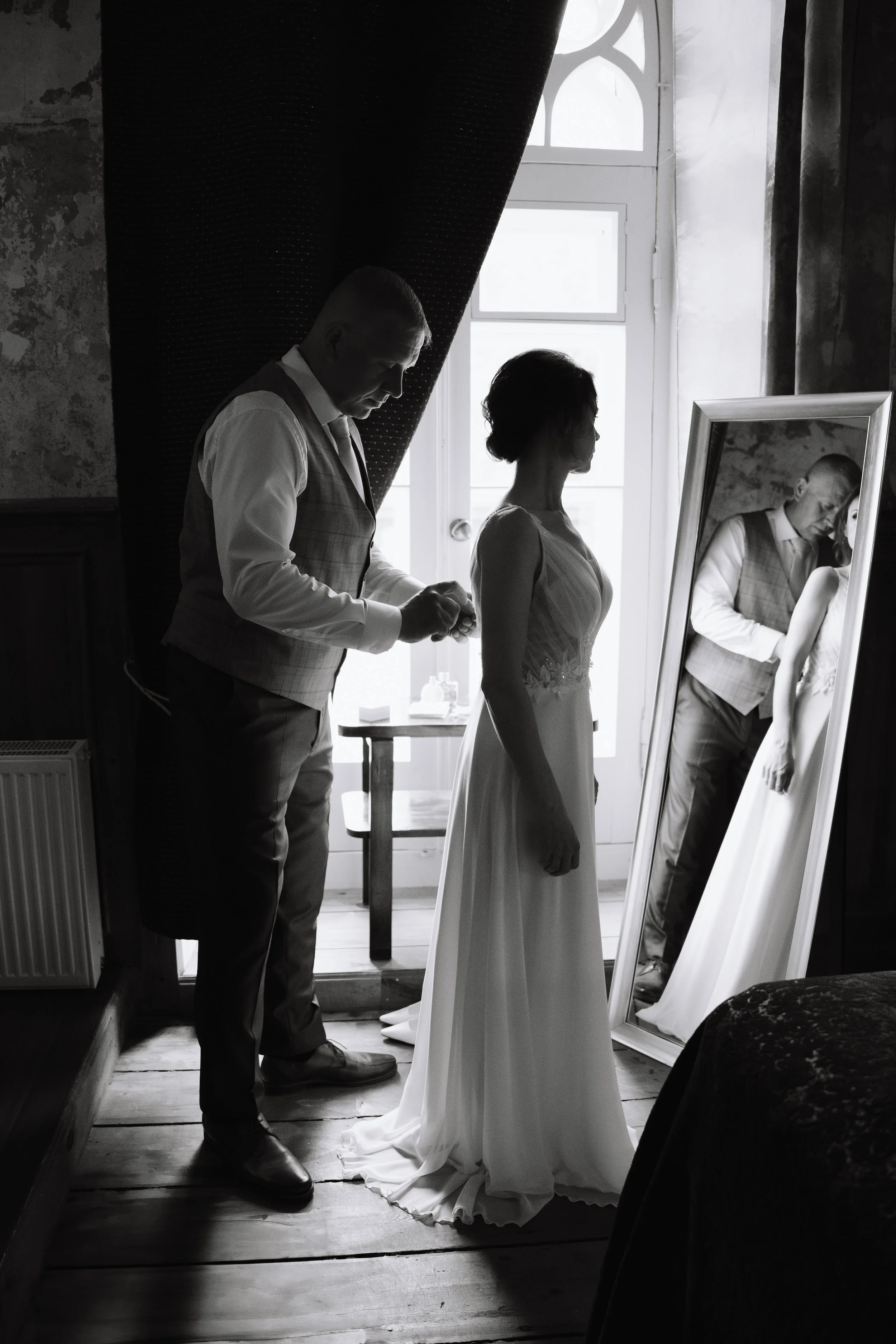 A bride standing in front of a mirror while a man, possibly her father, helps her with her wedding dress in a softly lit room.