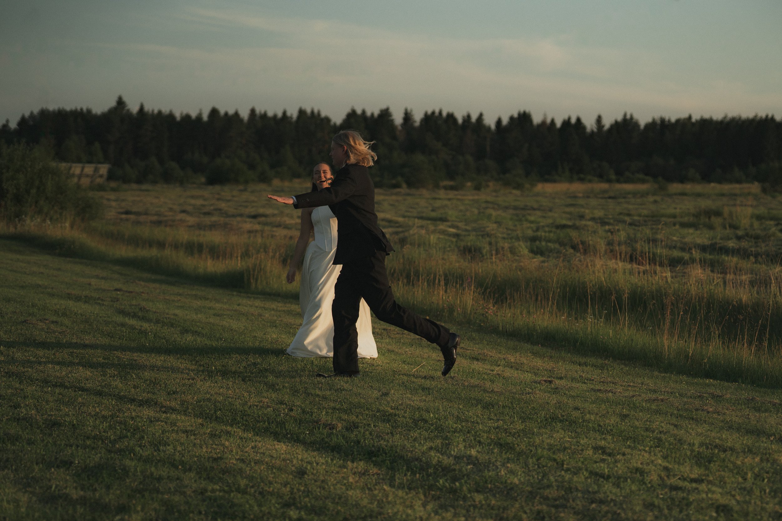 A man and woman dressed in wedding attire dancing on a grassy field at sunset, with trees in the background.