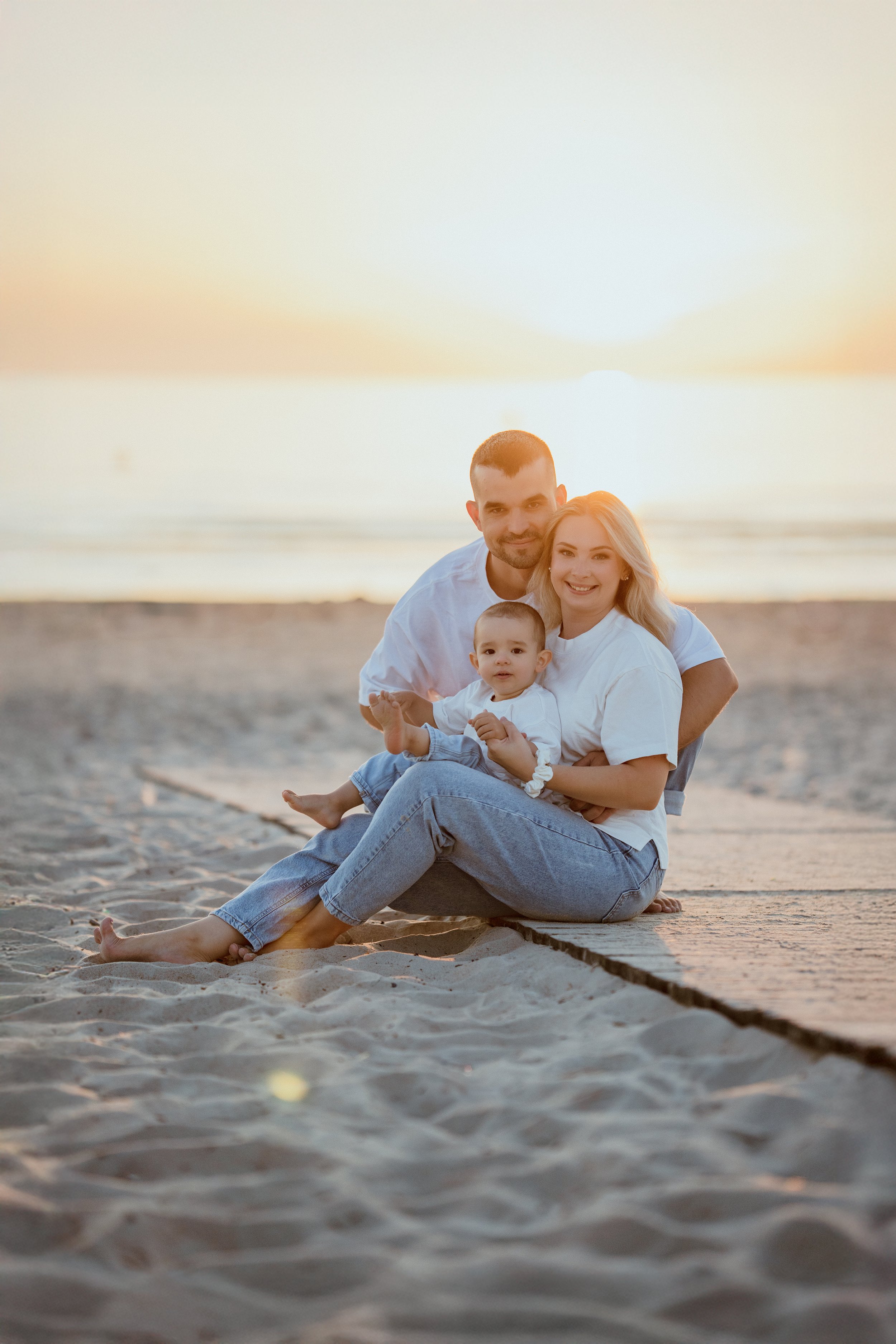 Family of three sitting on a beach during sunset, smiling at the camera.