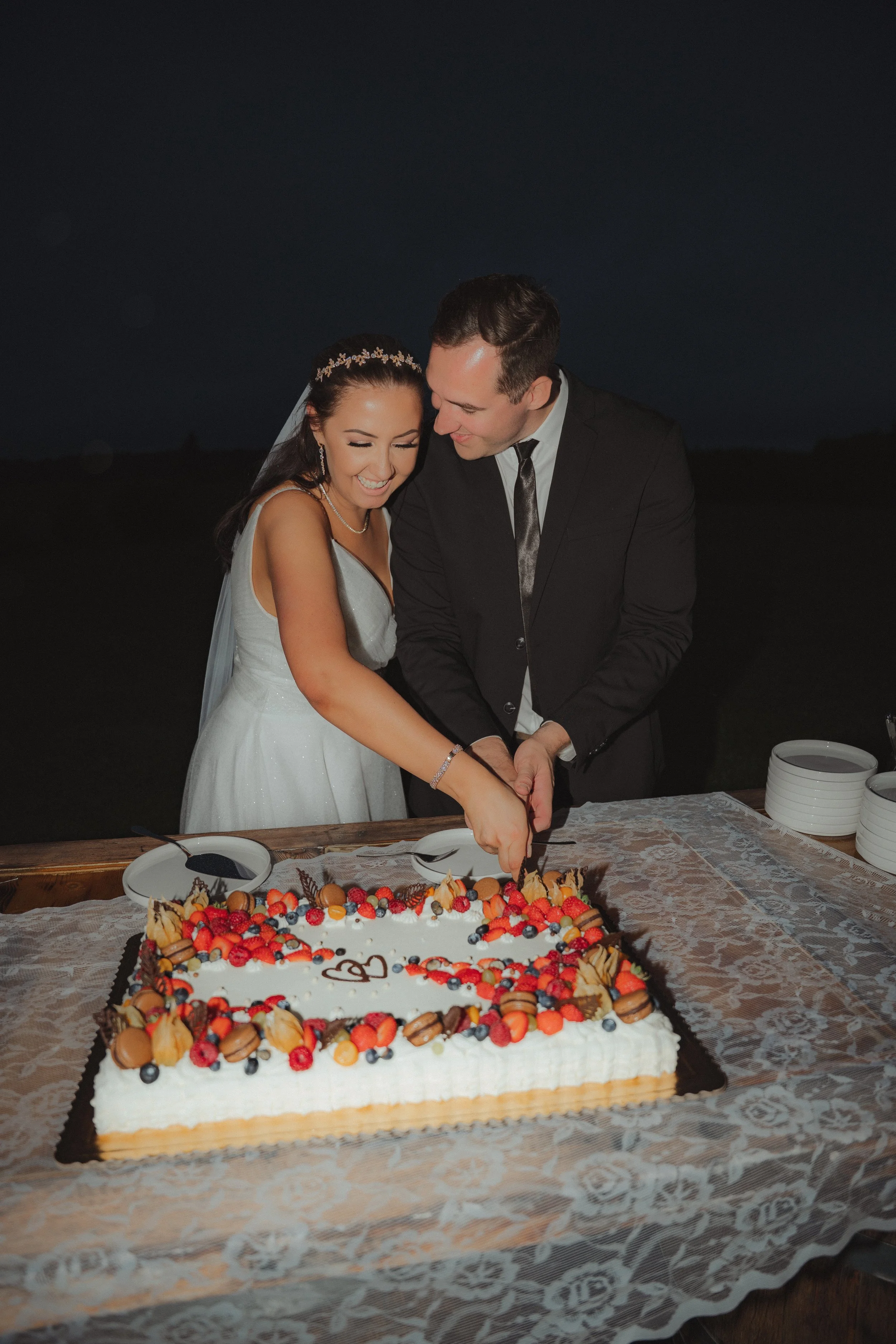 A bride and groom in wedding attire cut a large decorated wedding cake together at night.