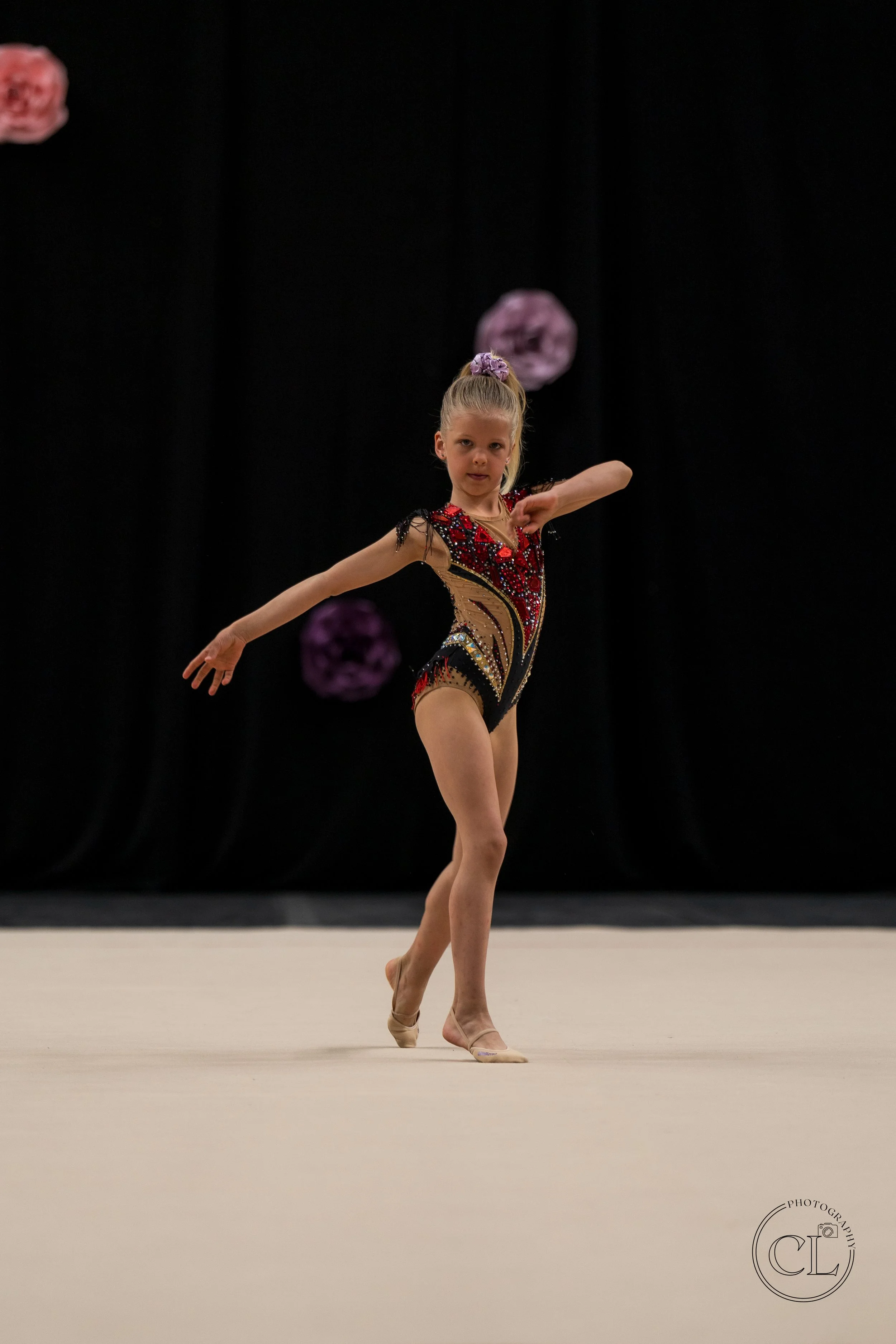 Young girl performing a gymnastics routine on a beige mat, wearing a colorful, decorated leotard, with black curtains and purple paper flowers in the background.
