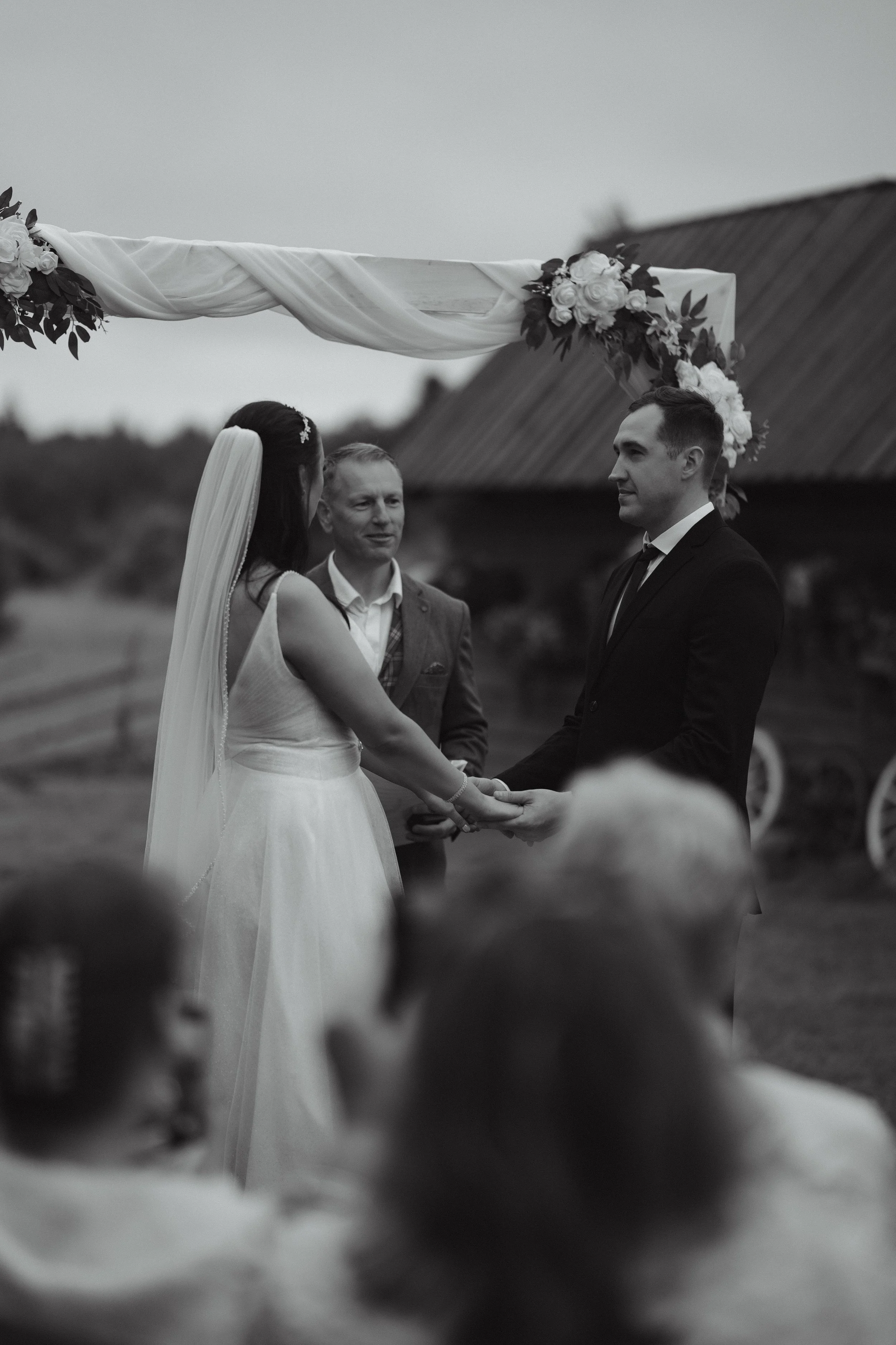 Black and white photo of a bride and groom exchanging vows during an outdoor wedding ceremony, with an officiant standing behind them under a decorated arch.