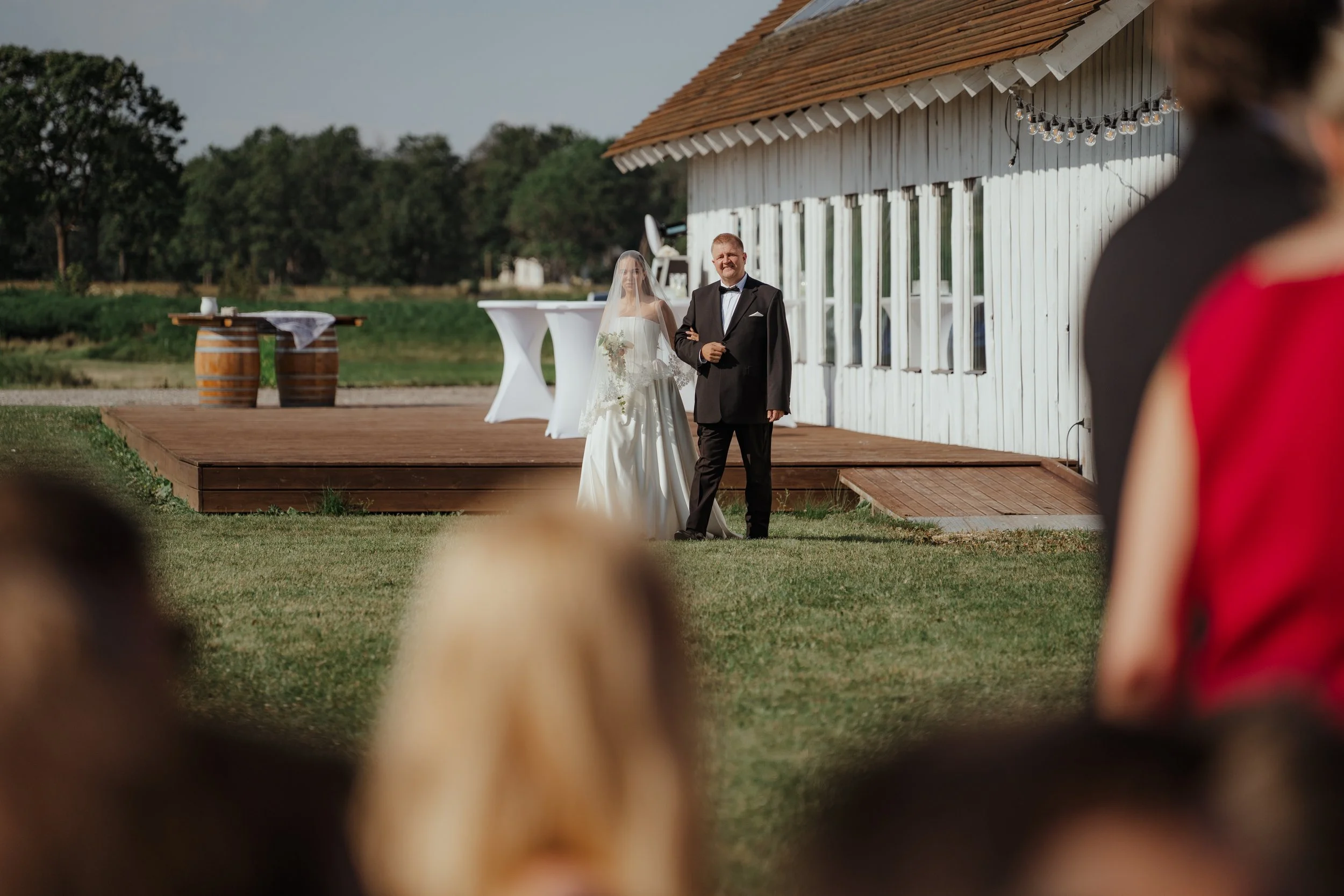 Bride walking down the aisle with her father at an outdoor wedding ceremony on a grassy lawn, near a white wooden building with a small stage and string lights in the background, as guests watch.