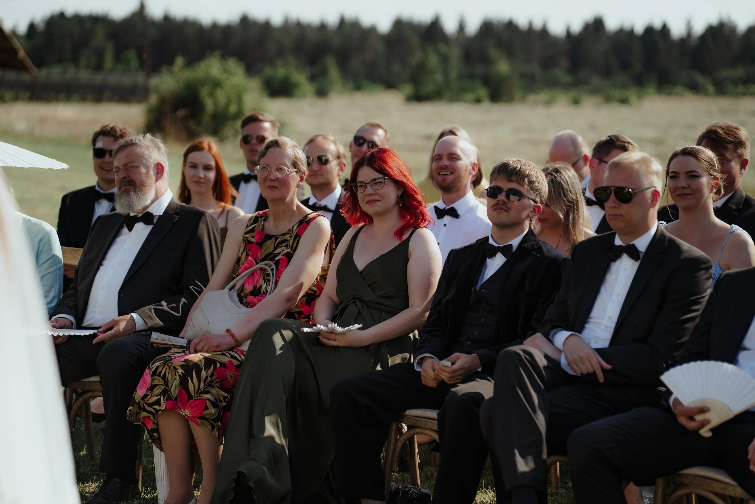 Group of people sitting outdoors in formal attire, attending a wedding ceremony on a sunny day.