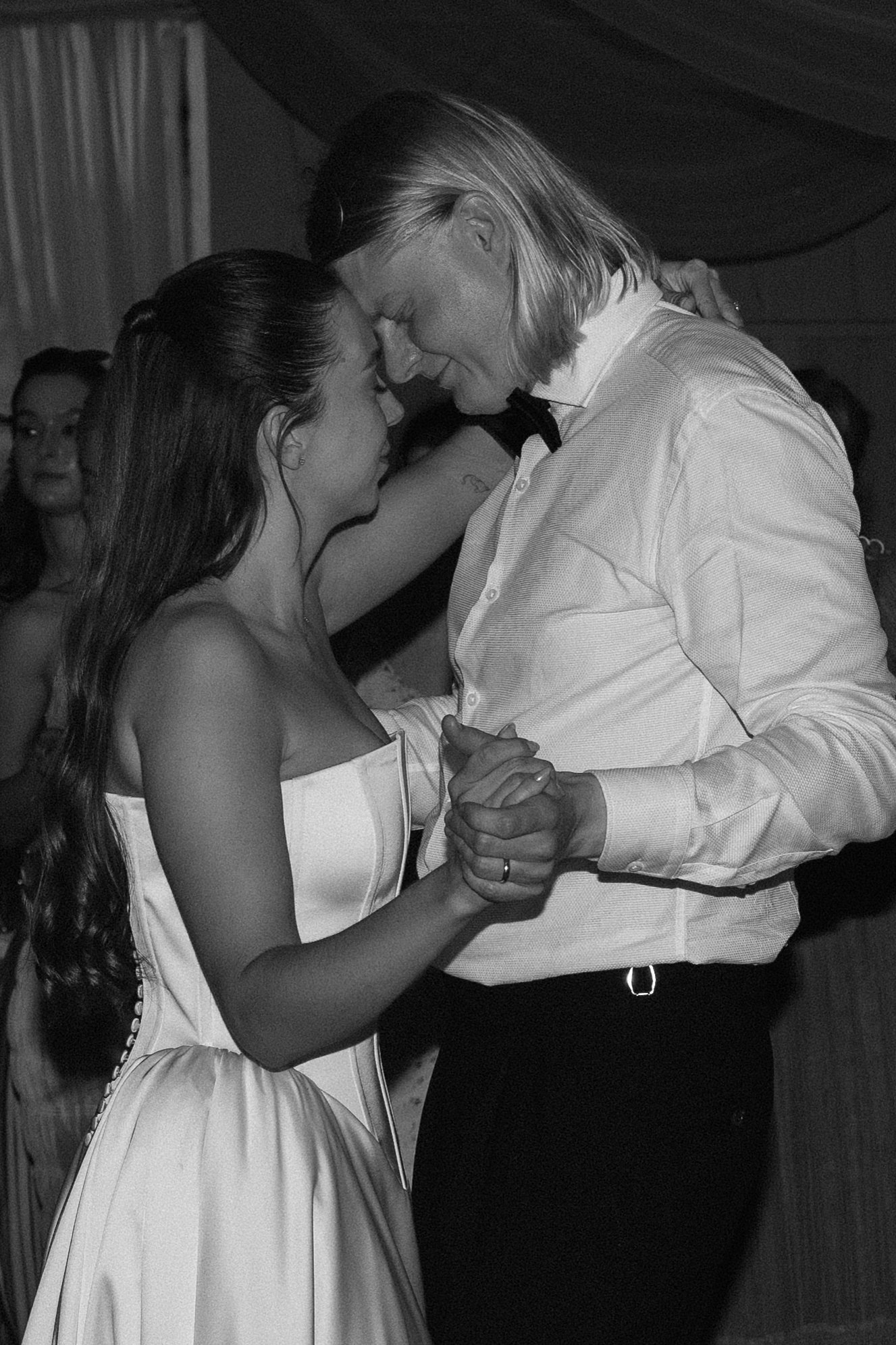 A black and white photo of a couple dancing closely, with foreheads touching, in an intimate moment at what seems to be a wedding or formal event.