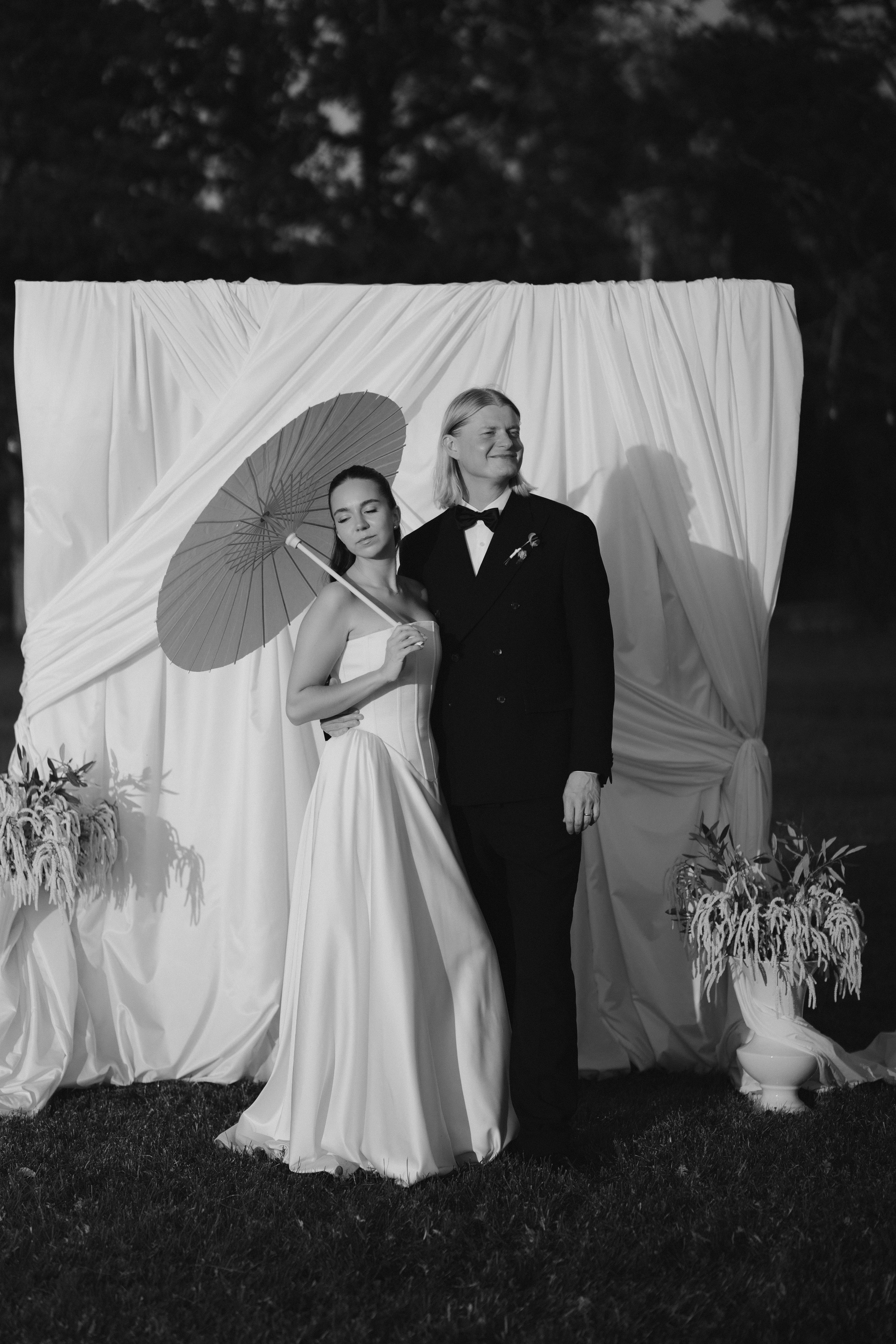 Black and white photo of a smiling man in a tuxedo and a woman in a strapless gown standing together outdoors, with a decorative backdrop and plants.