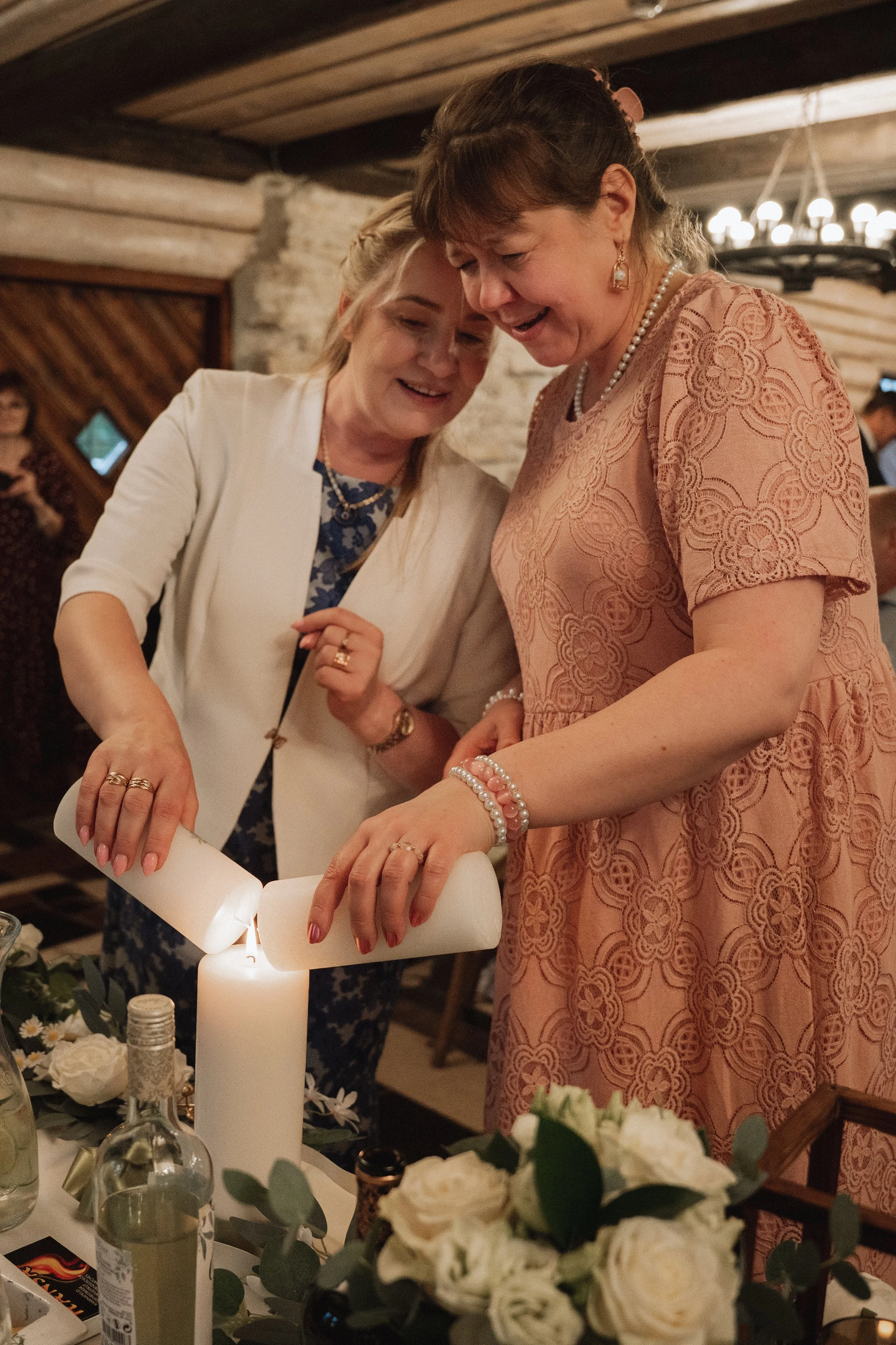 Two women lighting a candle together at a celebration or special event, smiling and wearing jewelry with flowers and bottles on the table.