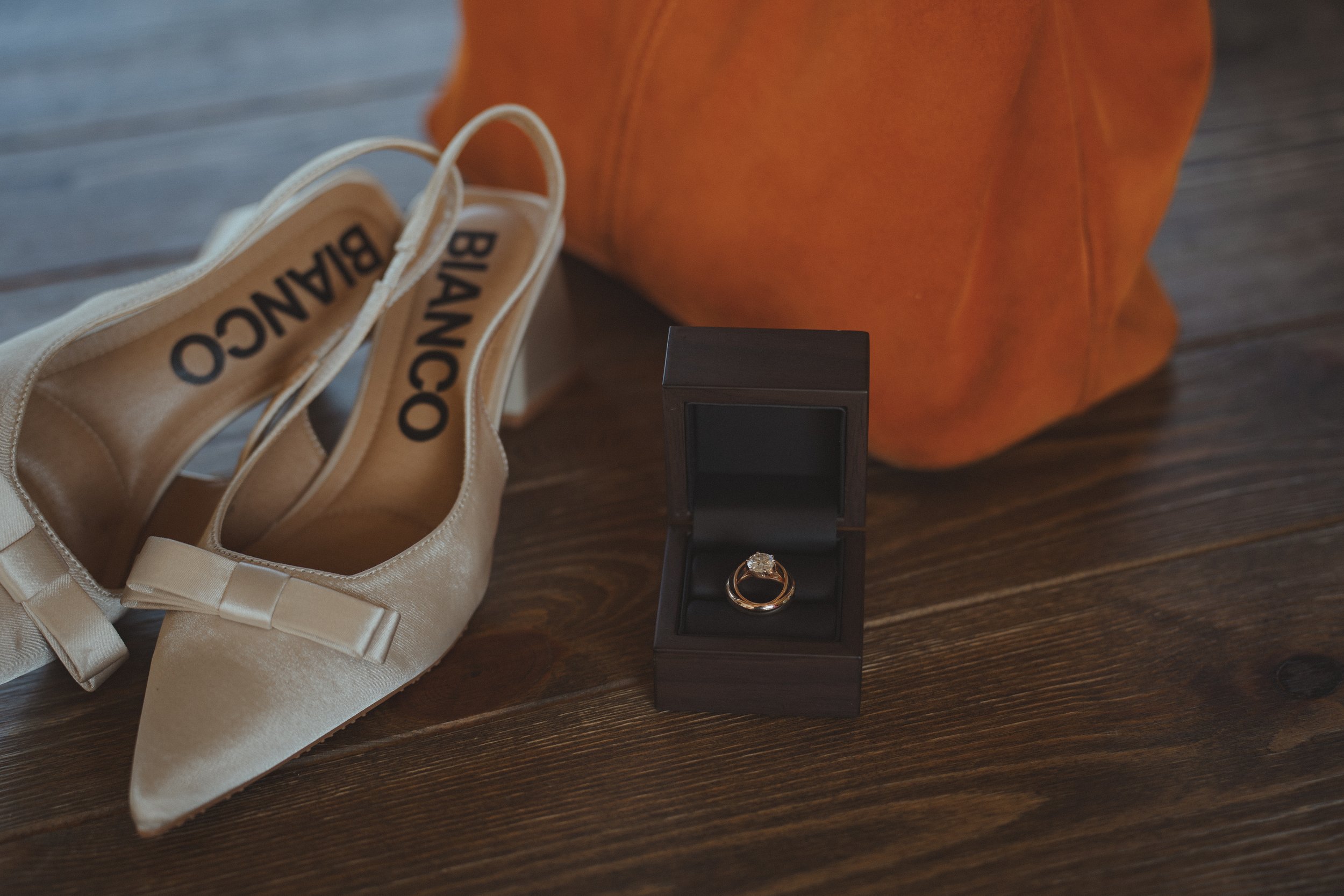 A pair of beige ballet flats with a bow, a black jewelry box containing a diamond engagement ring, an orange bag, and a wooden surface.