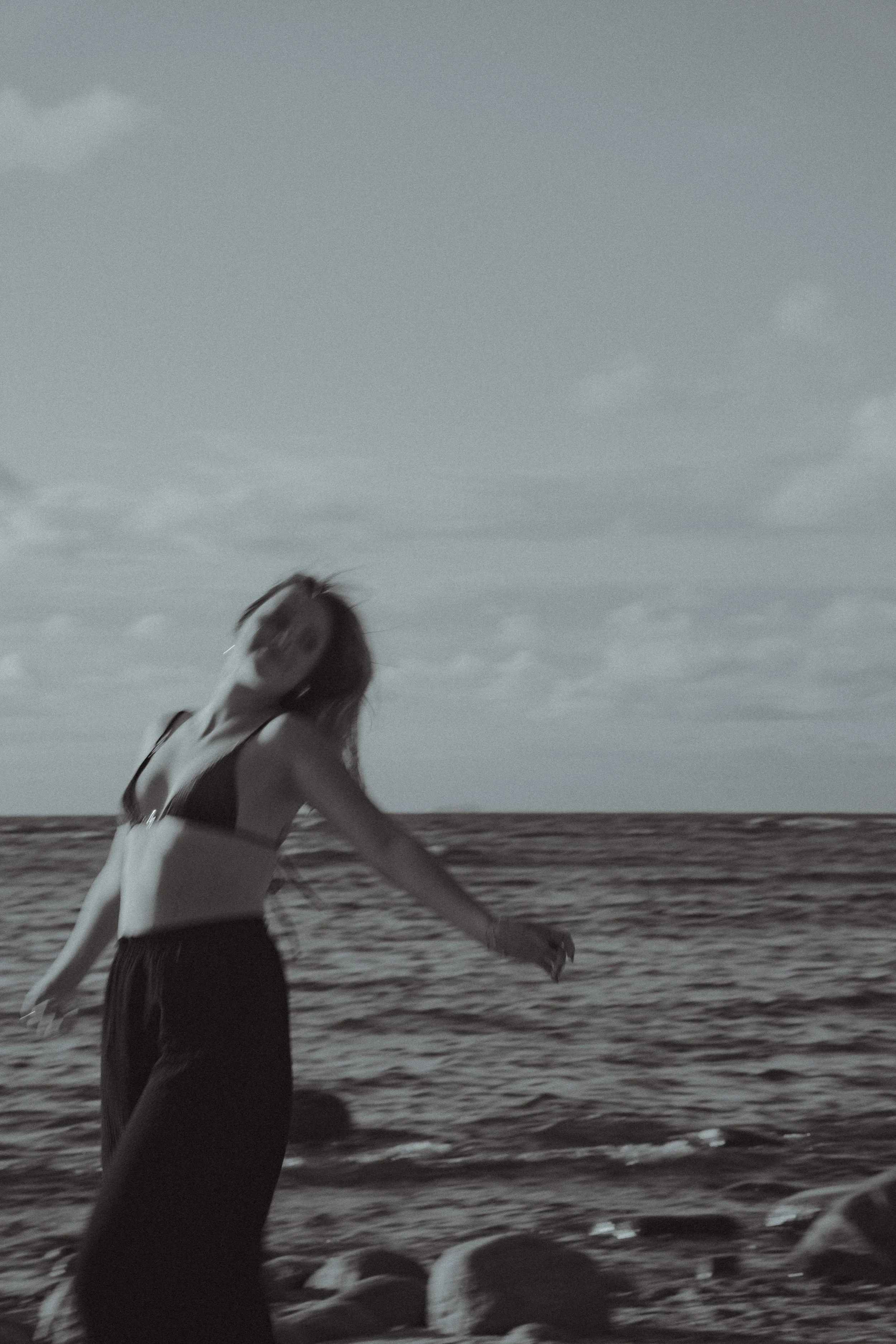 A woman with wavy hair in a dark swimsuit standing and dancing on a rocky beach by the ocean under cloudy sky.