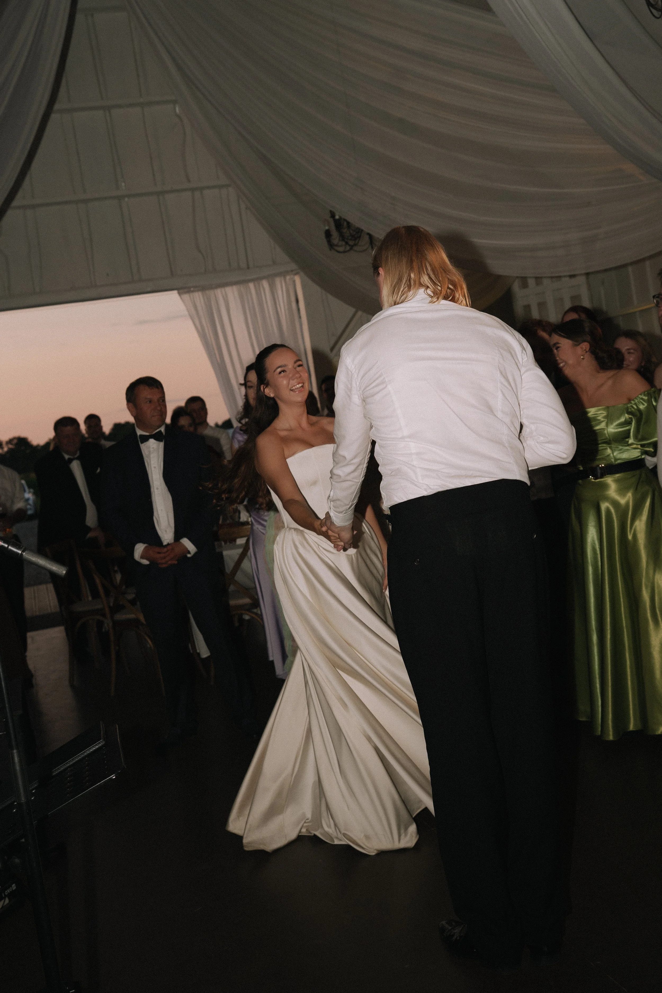 A bride and groom dancing at their wedding reception under a decorated tent with guests watching in the background.