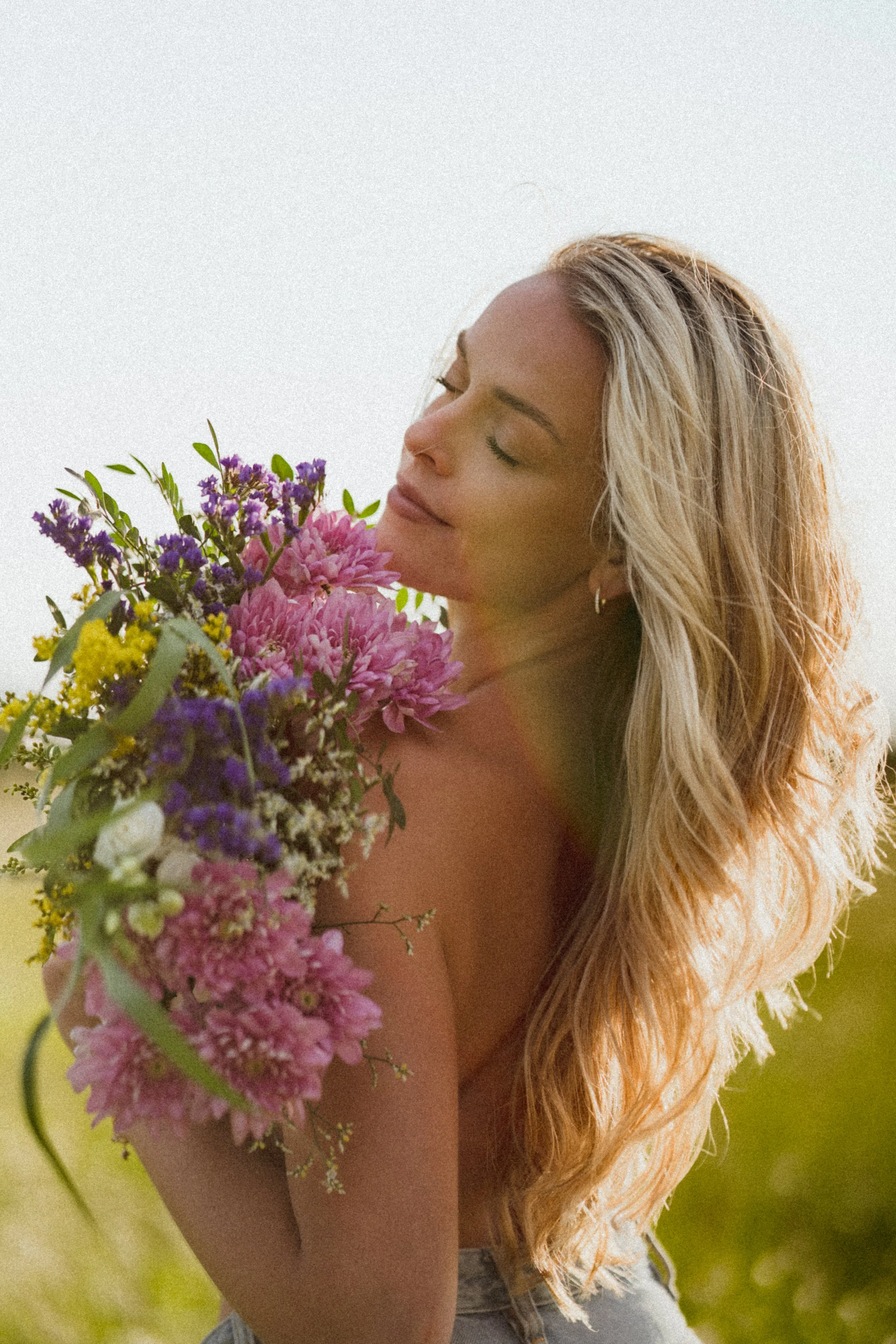 A woman with long, wavy blonde hair closed eyes holding a bouquet of colorful flowers, outdoors on a sunny day.