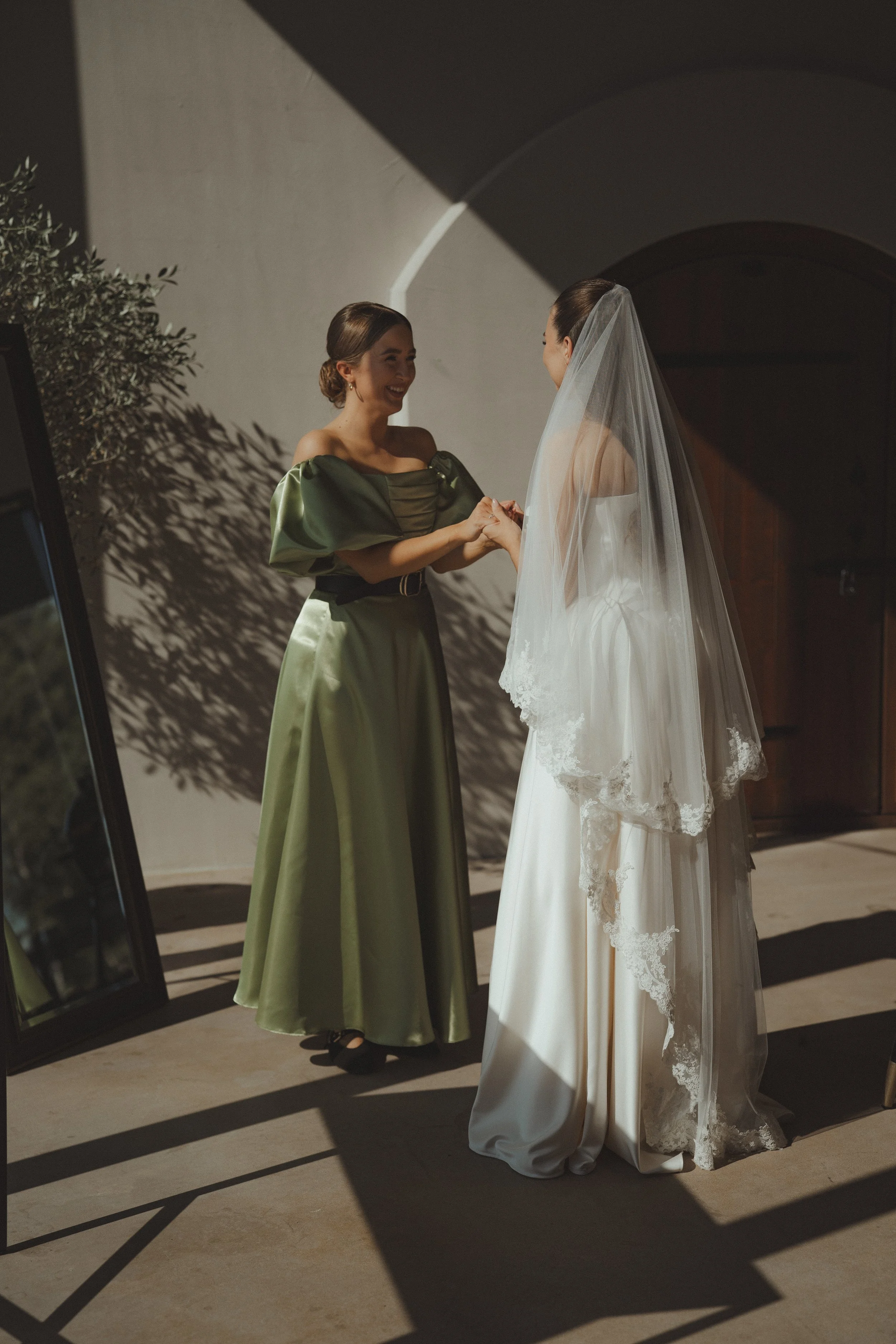 A bride in a white wedding dress with lace-trimmed veil holding hands with a woman in a green satin dress, smiling at each other indoors with sunlight casting shadows.