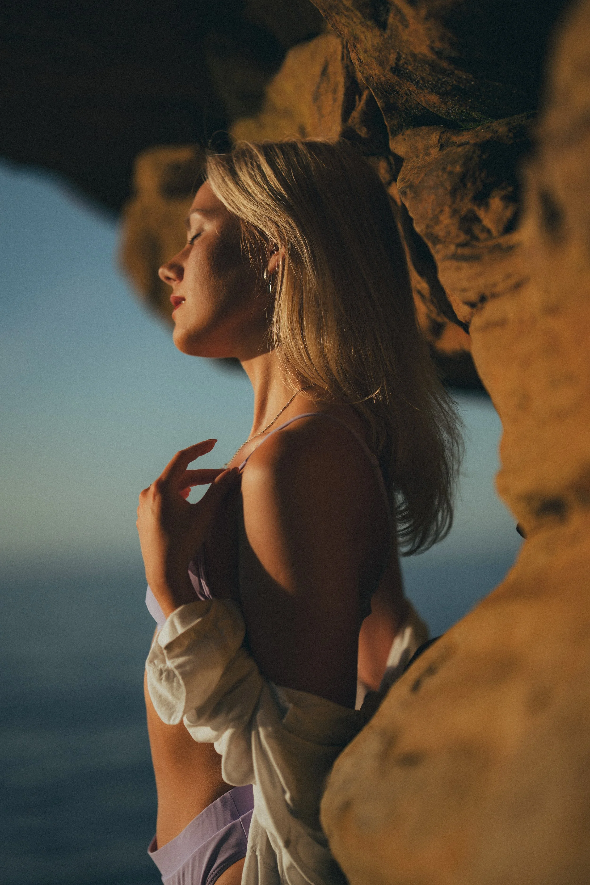 A woman with blonde hair and light skin standing with her eyes closed near a rocky formation at sunset or sunrise, with a calm body of water in the background.