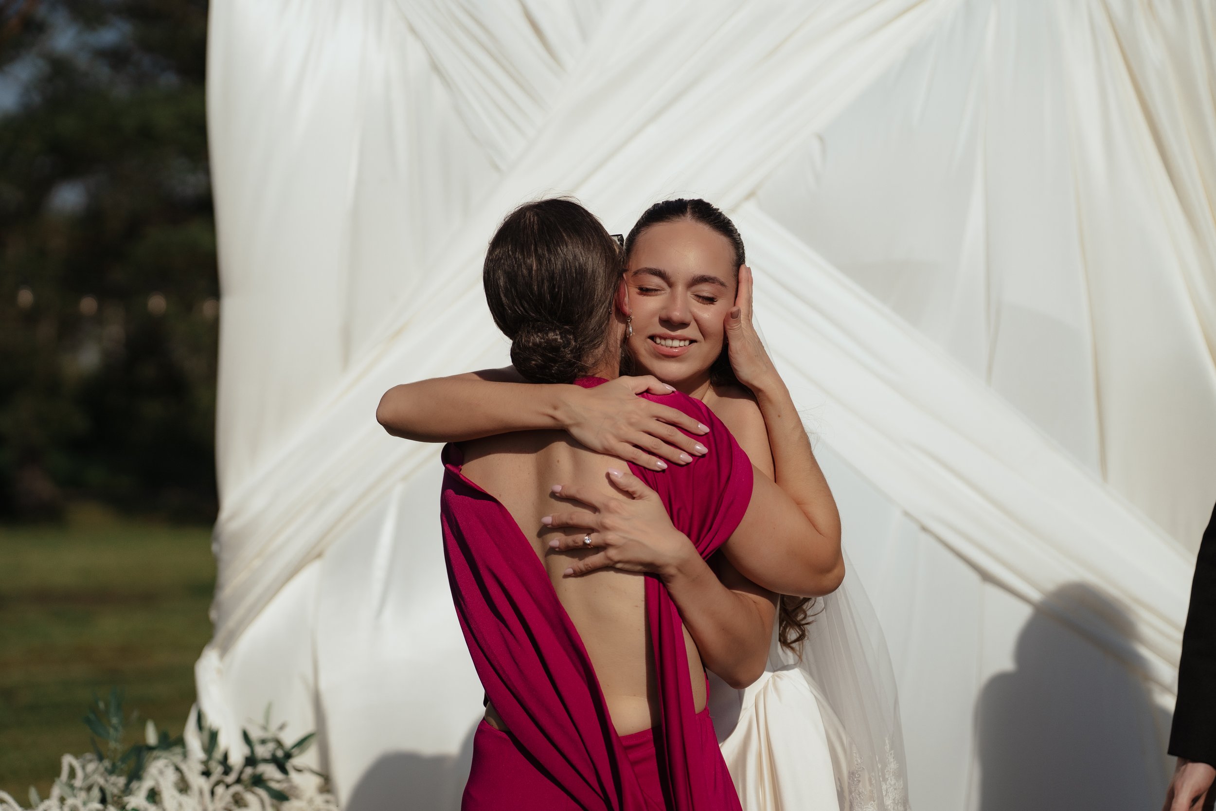 A woman in a wedding dress embraces an older woman in a red dress, both with eyes closed and emotional expressions, outdoors on a sunny day with white drapery in the background.