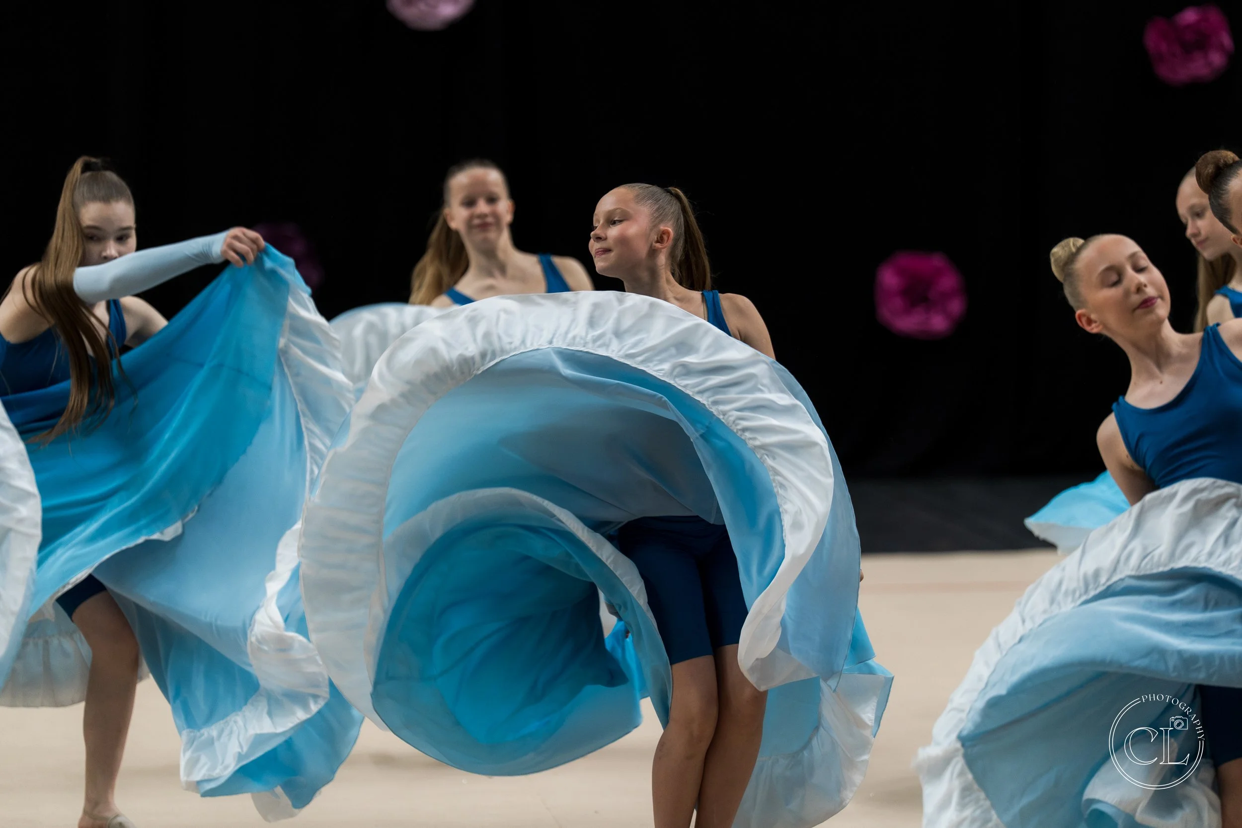 Young girls in blue dresses perform a dance with large, flowing blue and white fabric circles on stage with a black background and pink flower decorations.