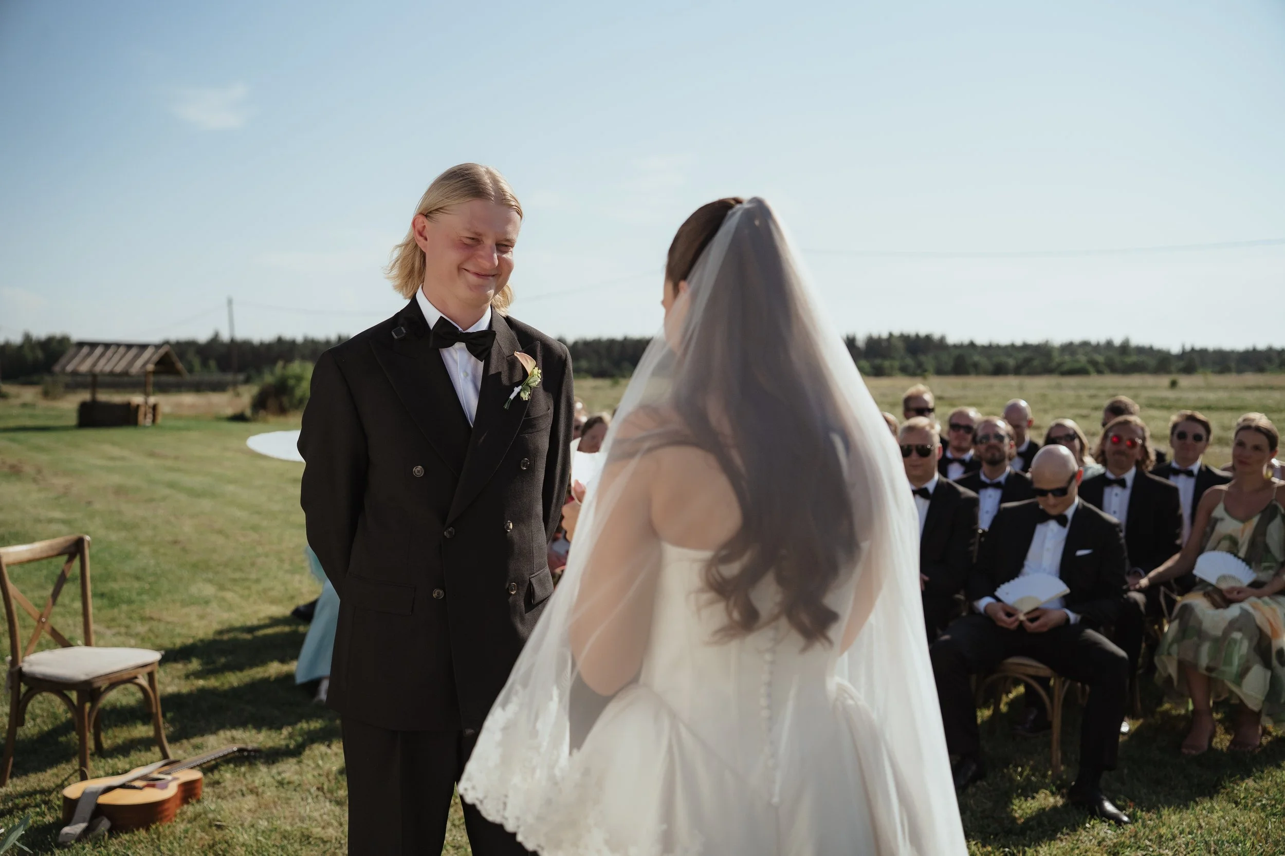 A wedding ceremony taking place outdoors in a grassy field with a man in a black tuxedo and a woman in a white wedding dress and veil, facing each other and smiling. Guests dressed in formal attire are seated in the background.