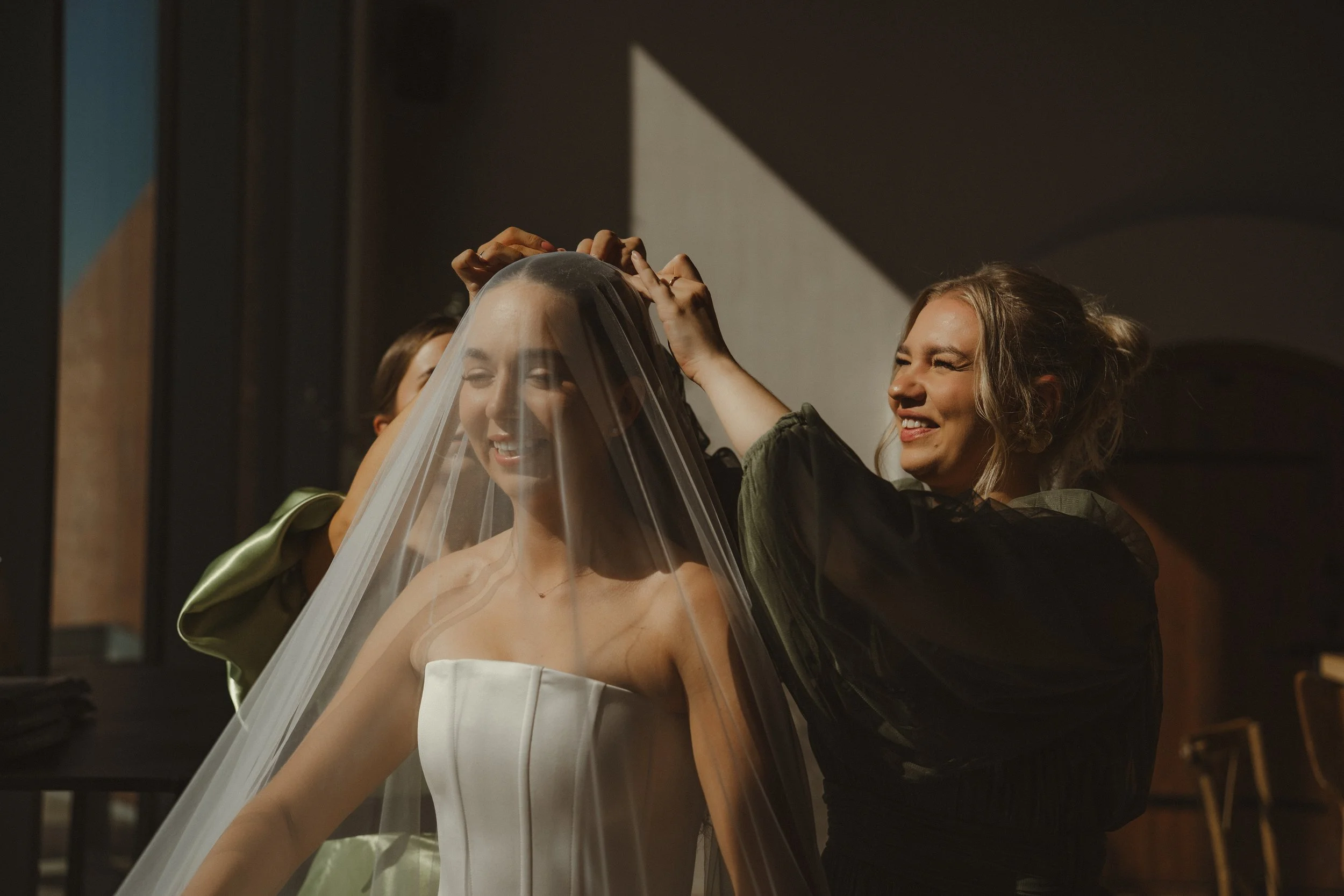 A bride getting ready with the help of a woman, who is adjusting her veil in a dimly lit room.
