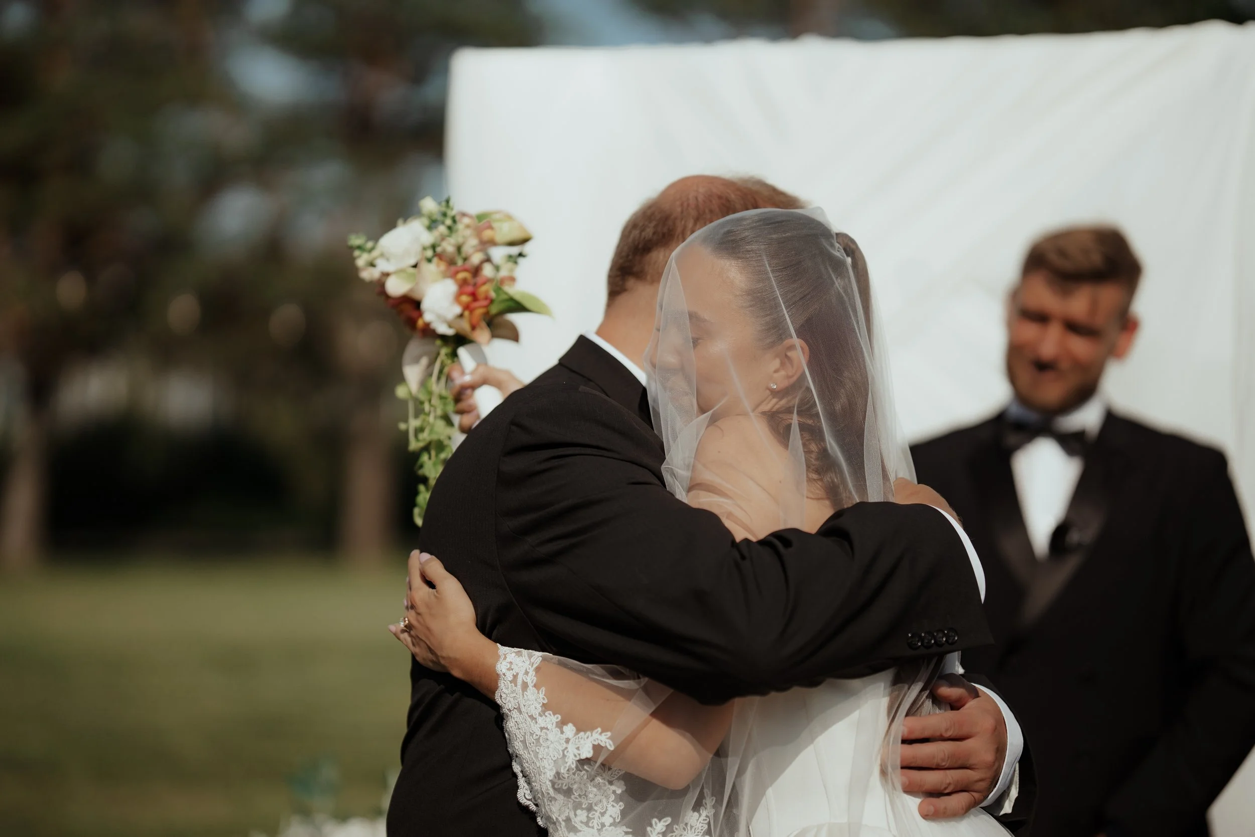 A bride and groom embracing during a wedding ceremony outdoors, with an officiant smiling in the background.