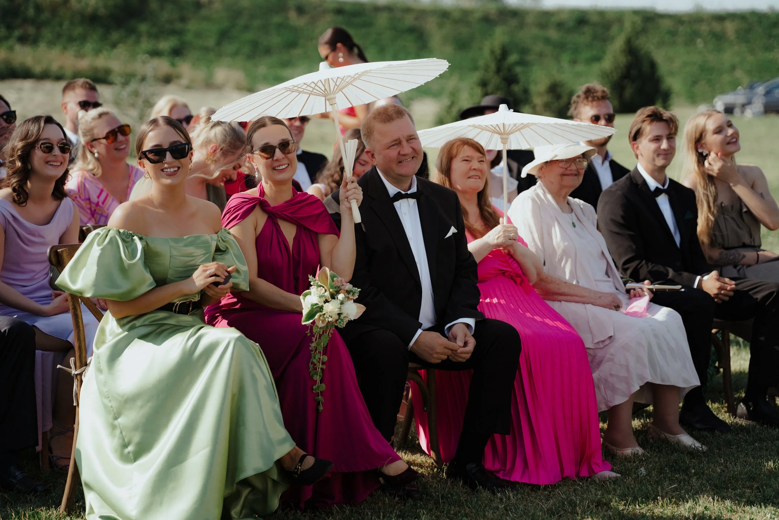 Group of people sitting outdoors at a wedding, some holding parasols, dressed in formal and colorful attire, smiling and enjoying the event.