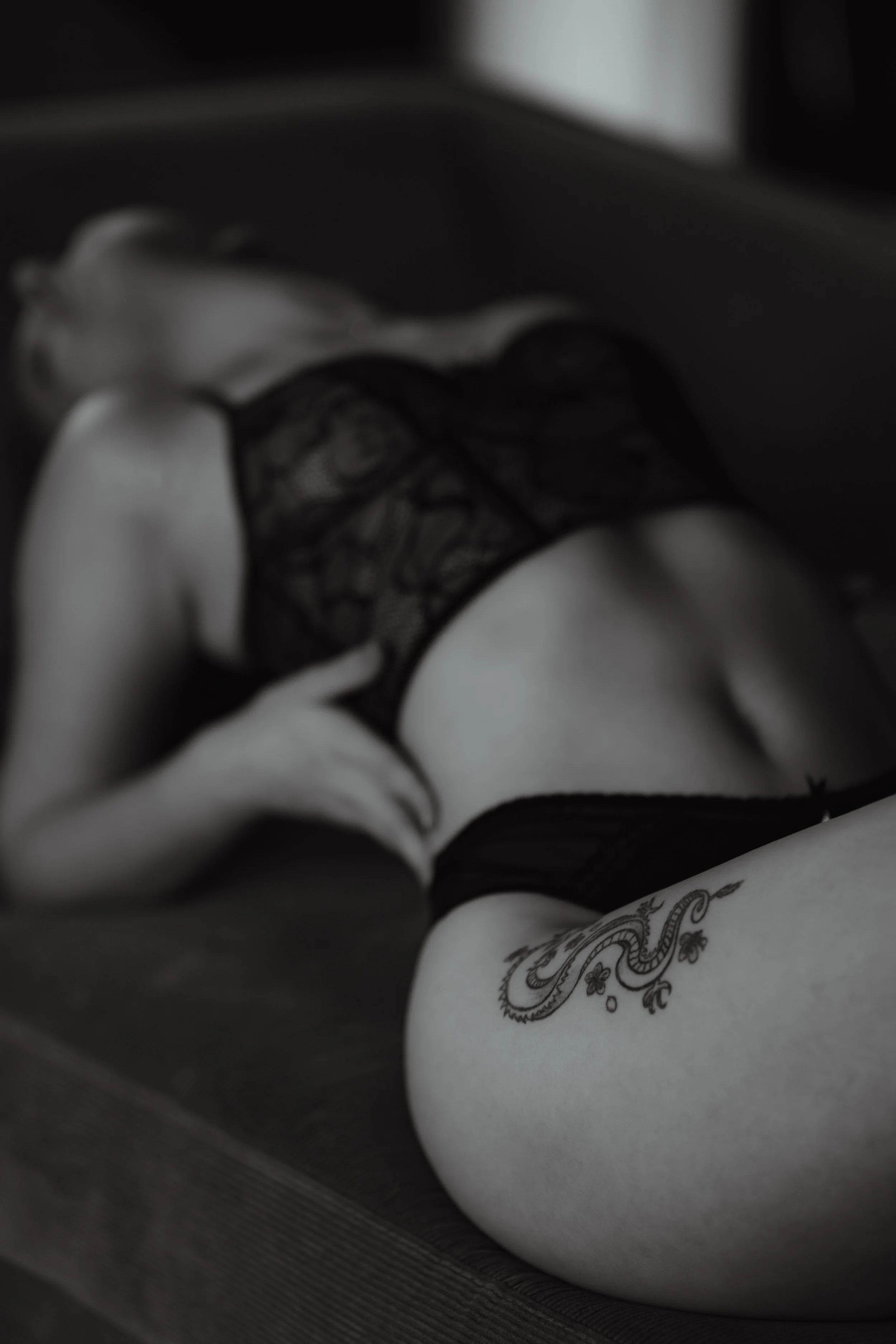 Black and white photo of a woman lying on her side on a flat surface, wearing lingerie with a tattoo on her thigh.