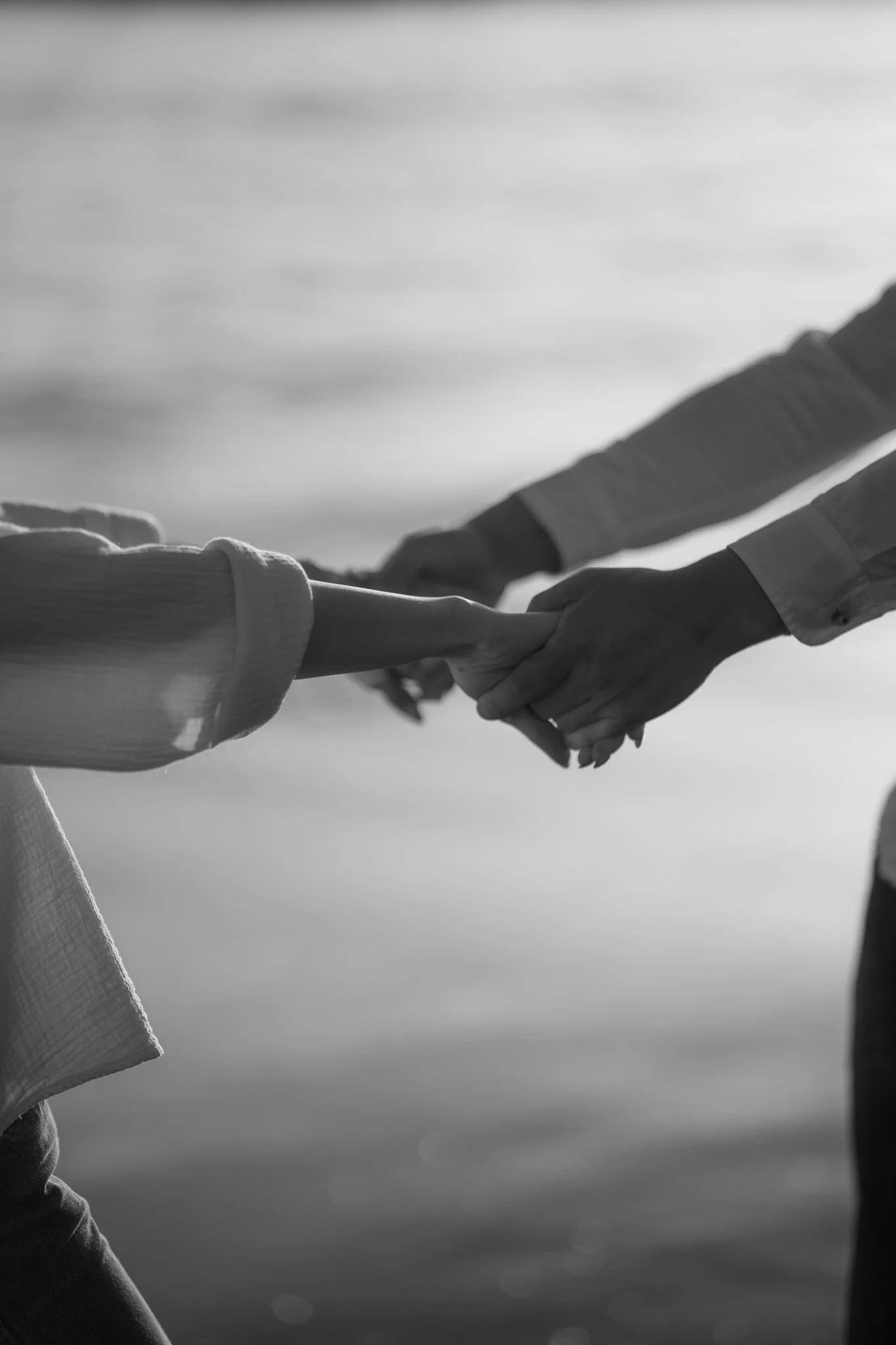 A black and white photo of two people holding hands, with one person's hand gently grasping the other's, against a blurred background of water.