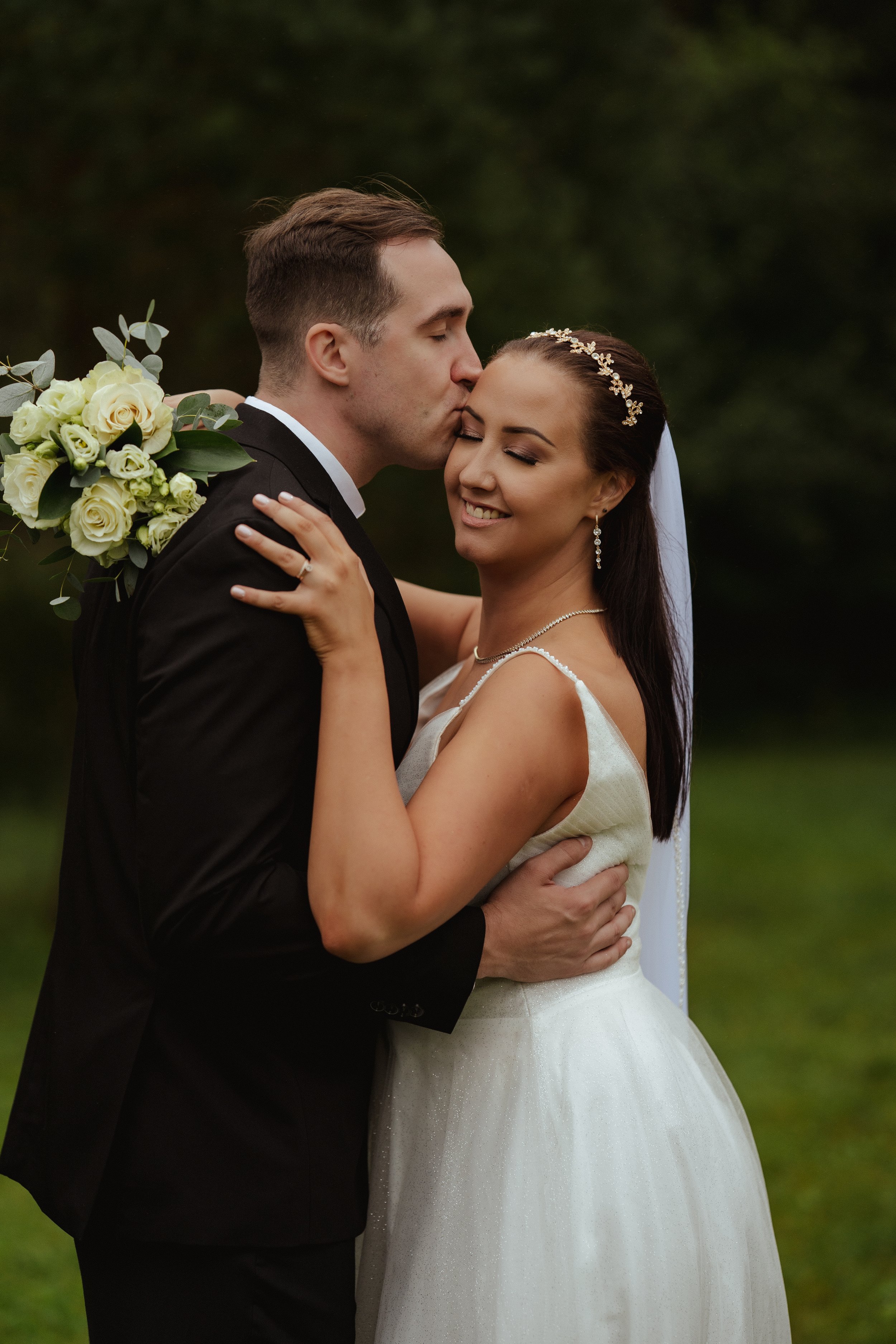 A newlywed couple is embracing outdoors, with the groom giving the bride a kiss on her forehead. The bride is smiling with eyes closed, holding a bouquet of white roses, and wearing a wedding dress with a sheer top and pearl accessories. The groom is