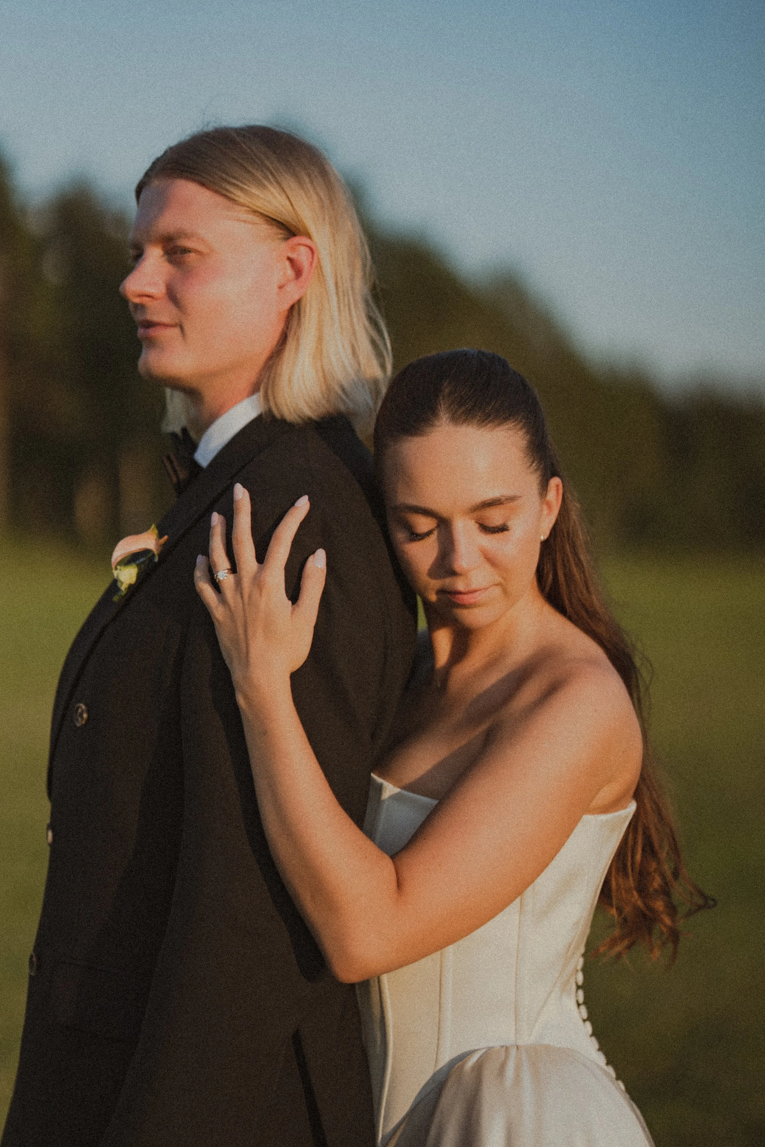 A couple, dressed in formal wedding attire, sharing a tender moment outdoors during sunset. The woman is embracing the man from behind, with her eyes closed and a serene expression.