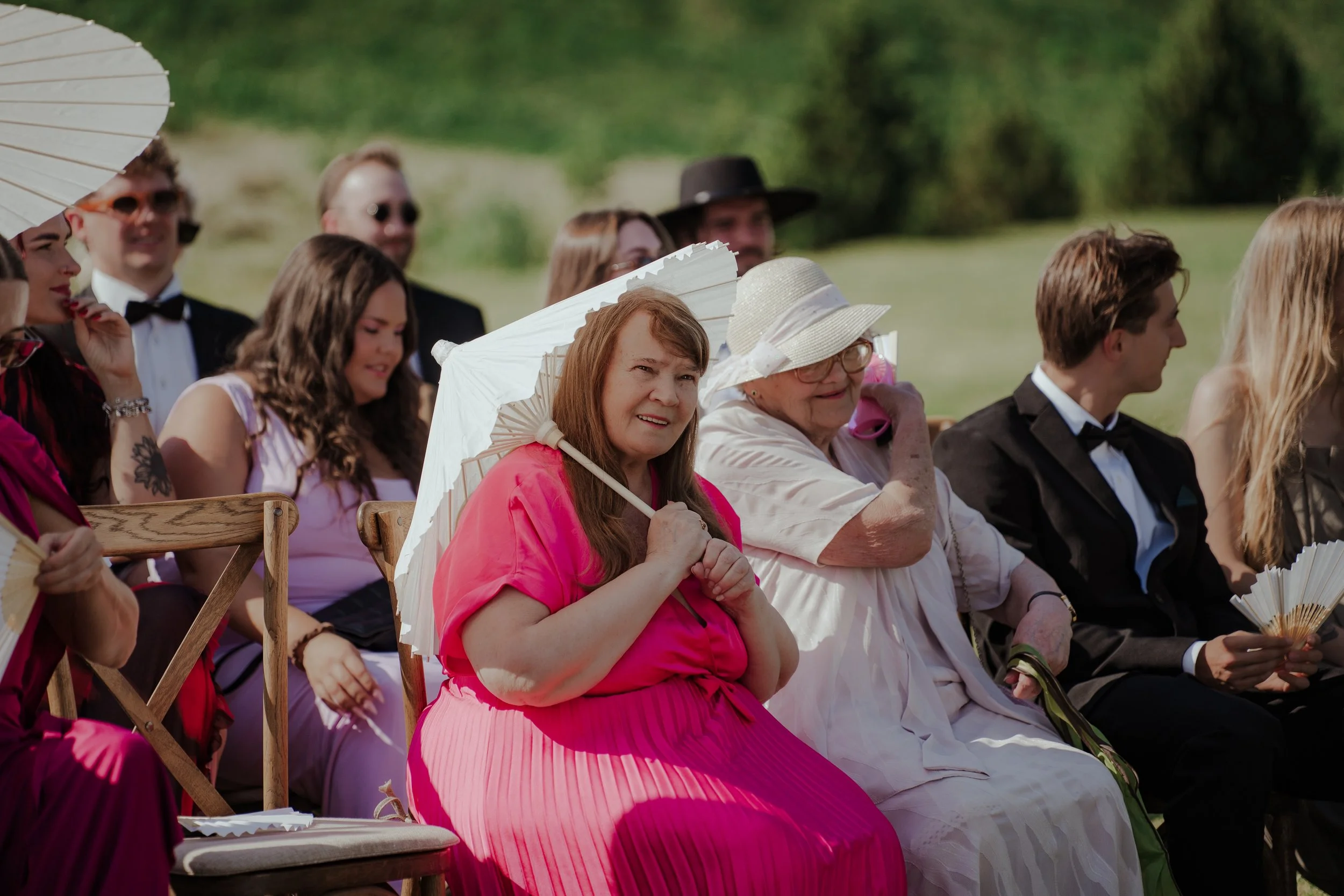 A group of people dressed in formal attire sitting outdoors during a sunny day, some holding umbrellas for shade.