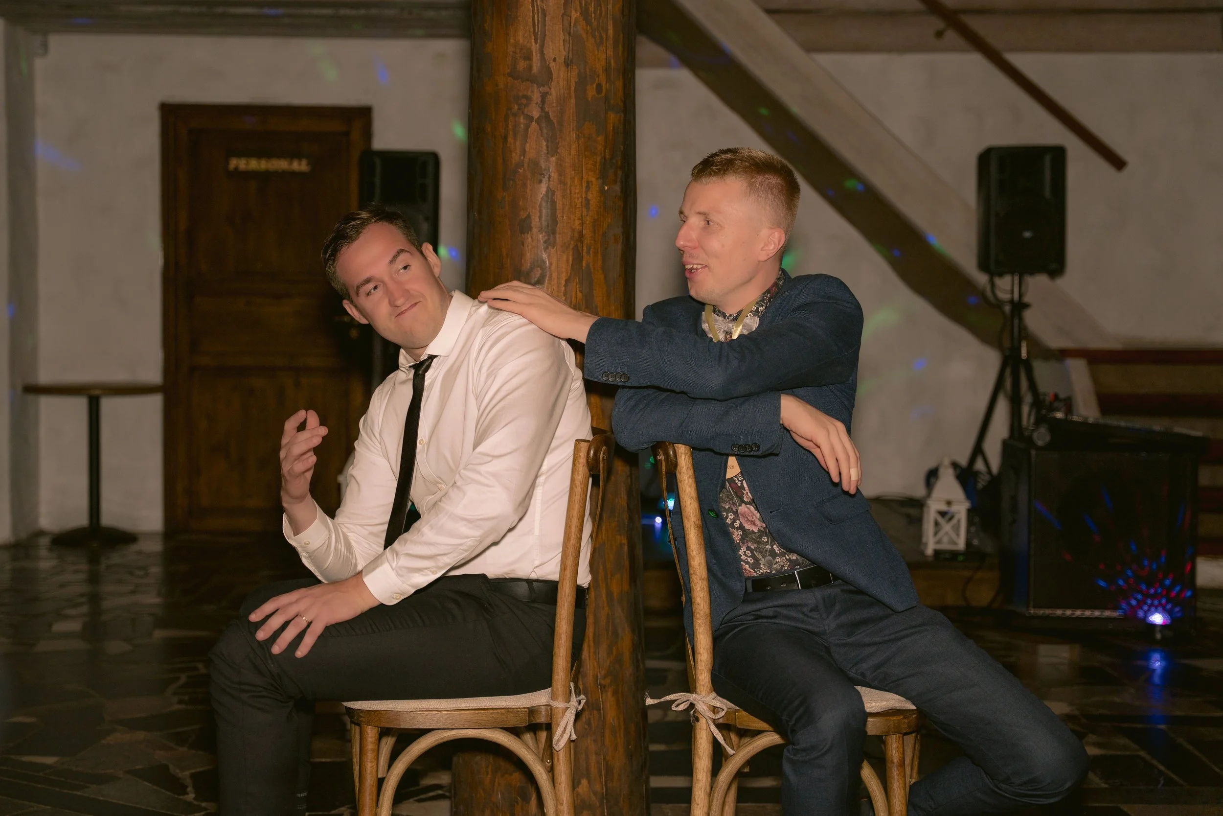 Two men are sitting on wooden chairs back-to-back, pretend fighting or joking with each other at an indoor event, with one man placing his hand on the other's shoulder.