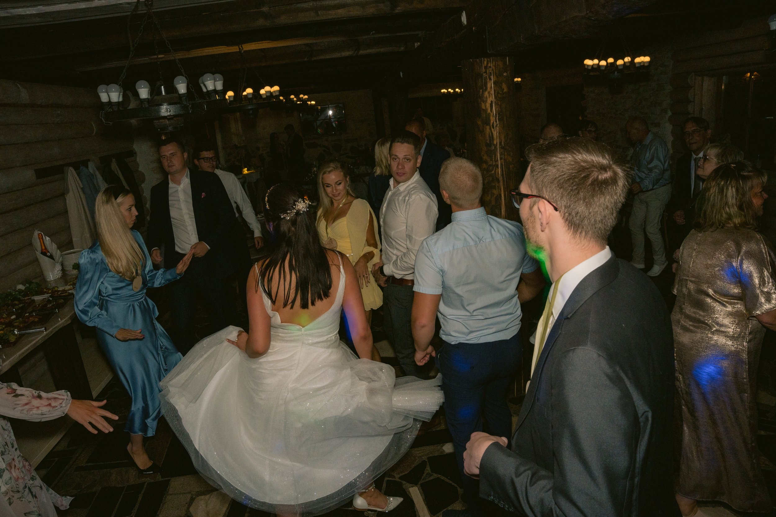 People dancing and socializing at a wedding reception in a rustic venue with wooden walls and low lighting.