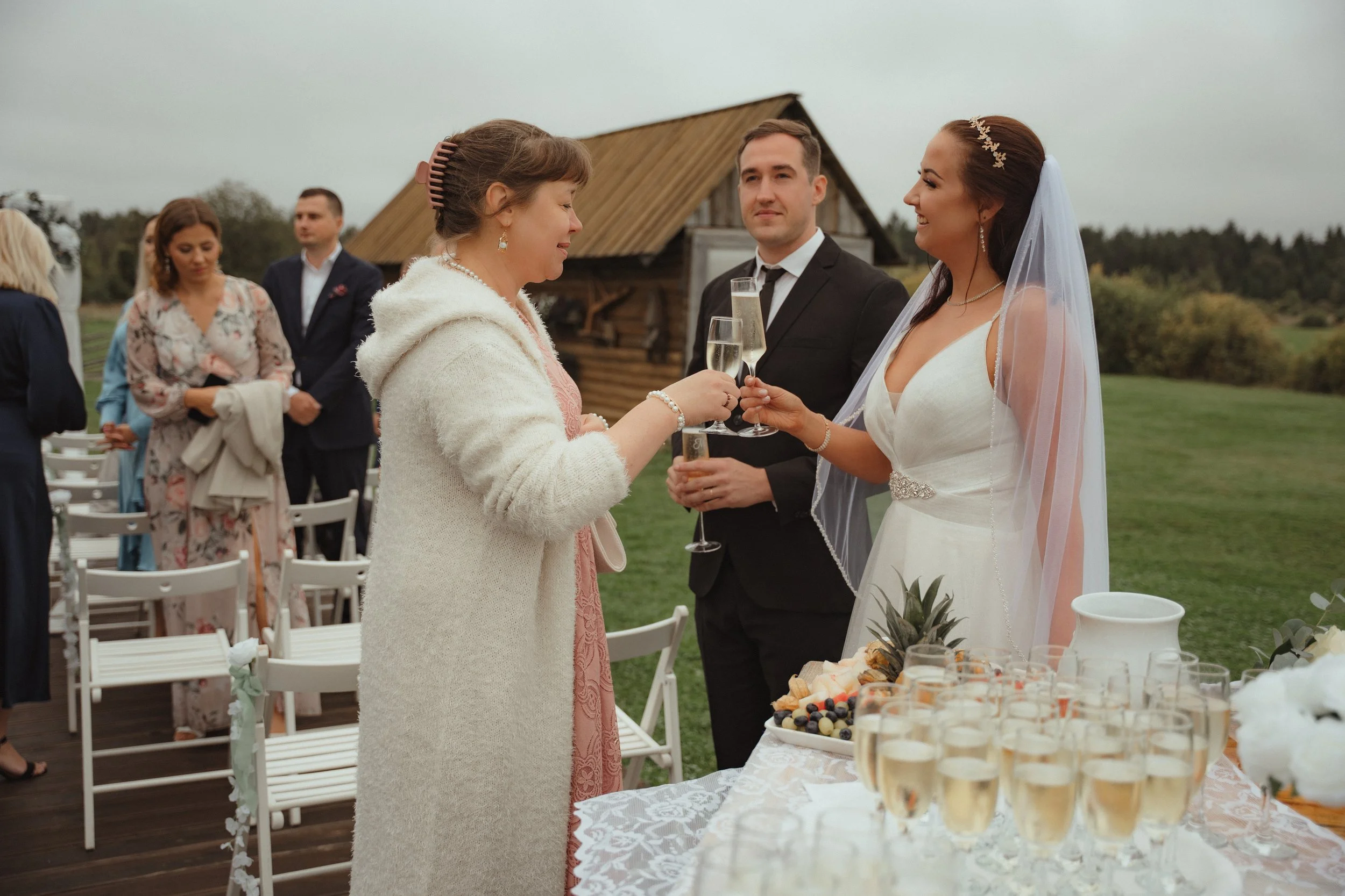 Bride and a woman toasting with champagne glasses at an outdoor wedding reception with guests in the background and a rustic wooden cabin.