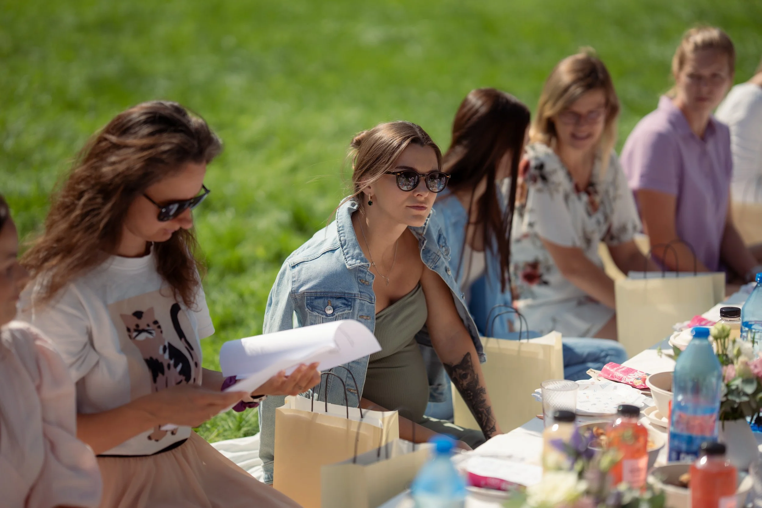 Group of women sitting outdoors at a table with food, drinks, and gift bags, engaging in conversation on a sunny day.
