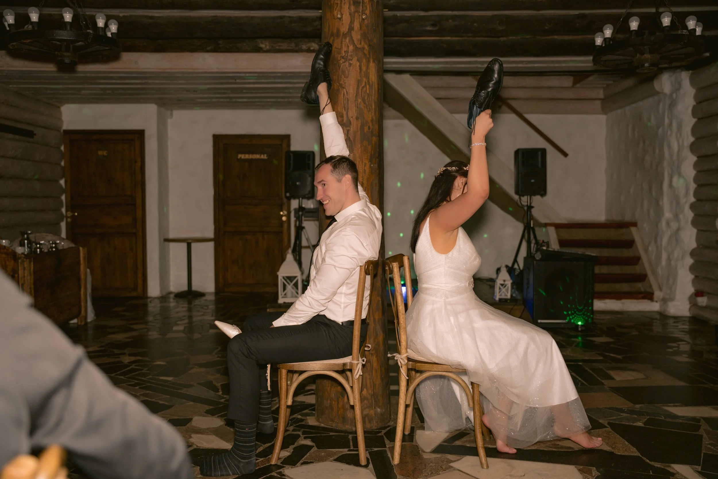 A bride and groom are sitting back-to-back on chairs, with a wooden pole between them, playing a game at their wedding reception. The groom is smiling, sitting on a chair facing the camera, and the bride is also smiling, with one arm raised holding a