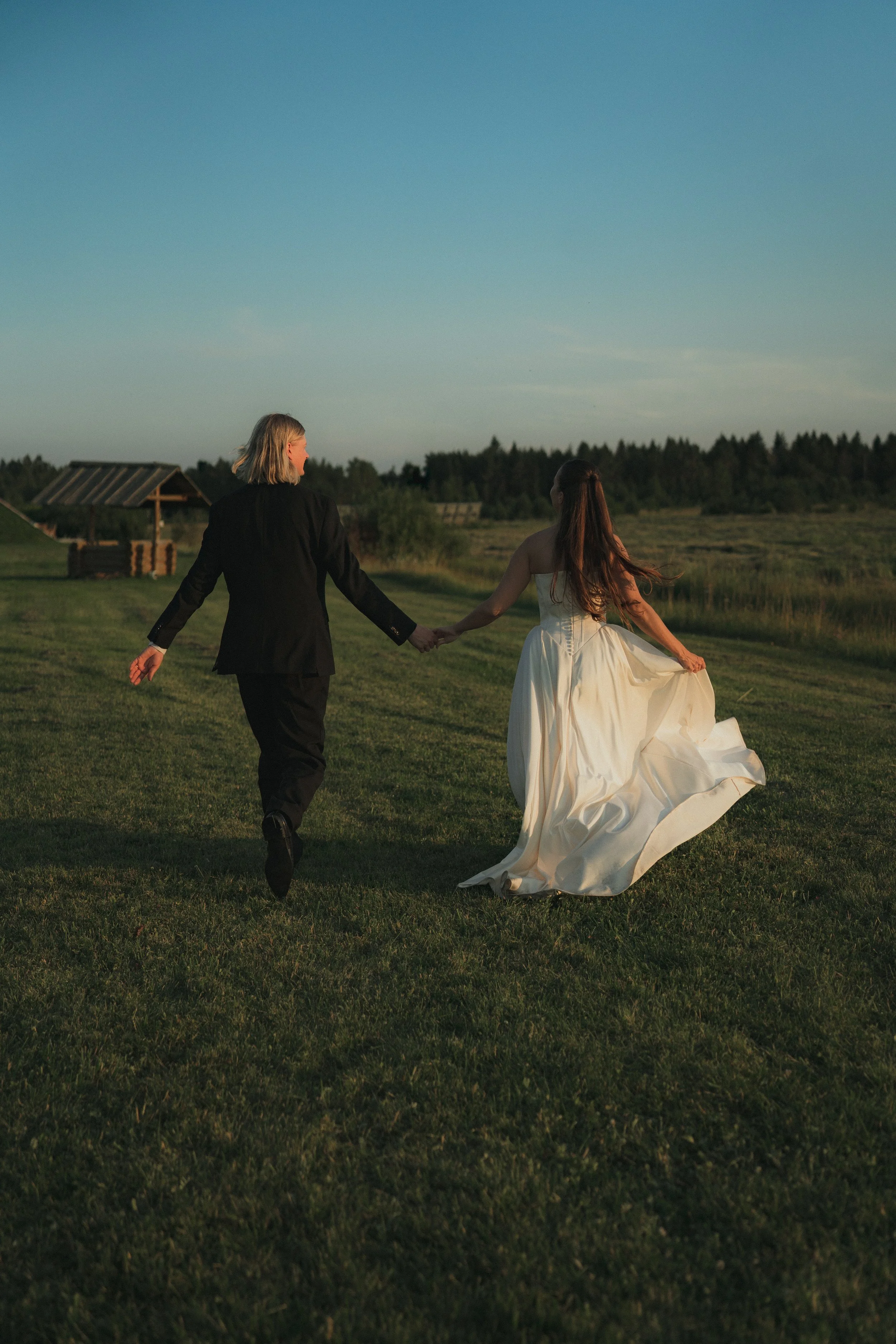 A couple walking hand in hand across a grassy field during sunset, with the woman wearing a white wedding dress and the man in a black suit.