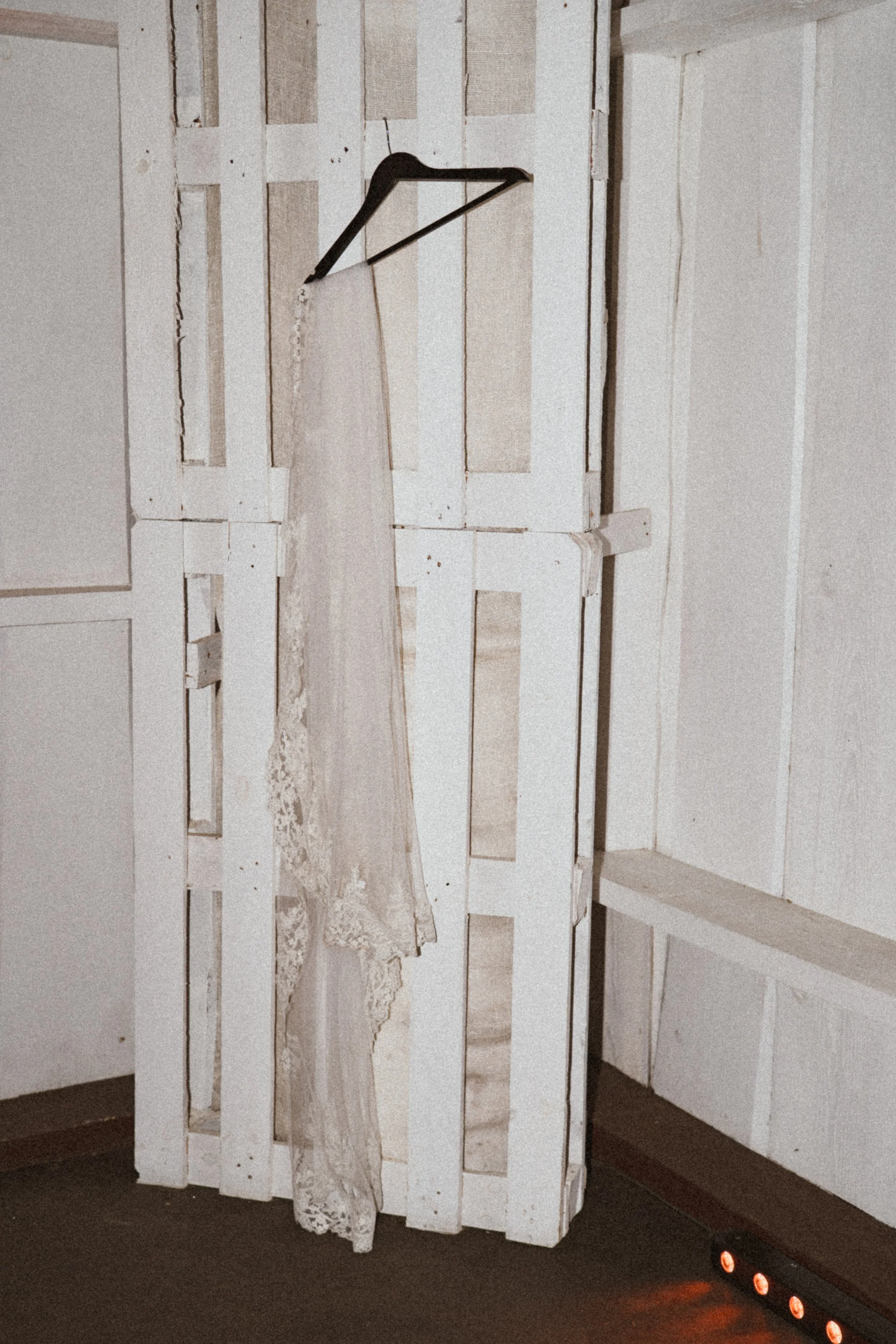 A wedding dress hangs on a black hanger against a white wooden pallet backdrop, with warm lighting and a dark floor.