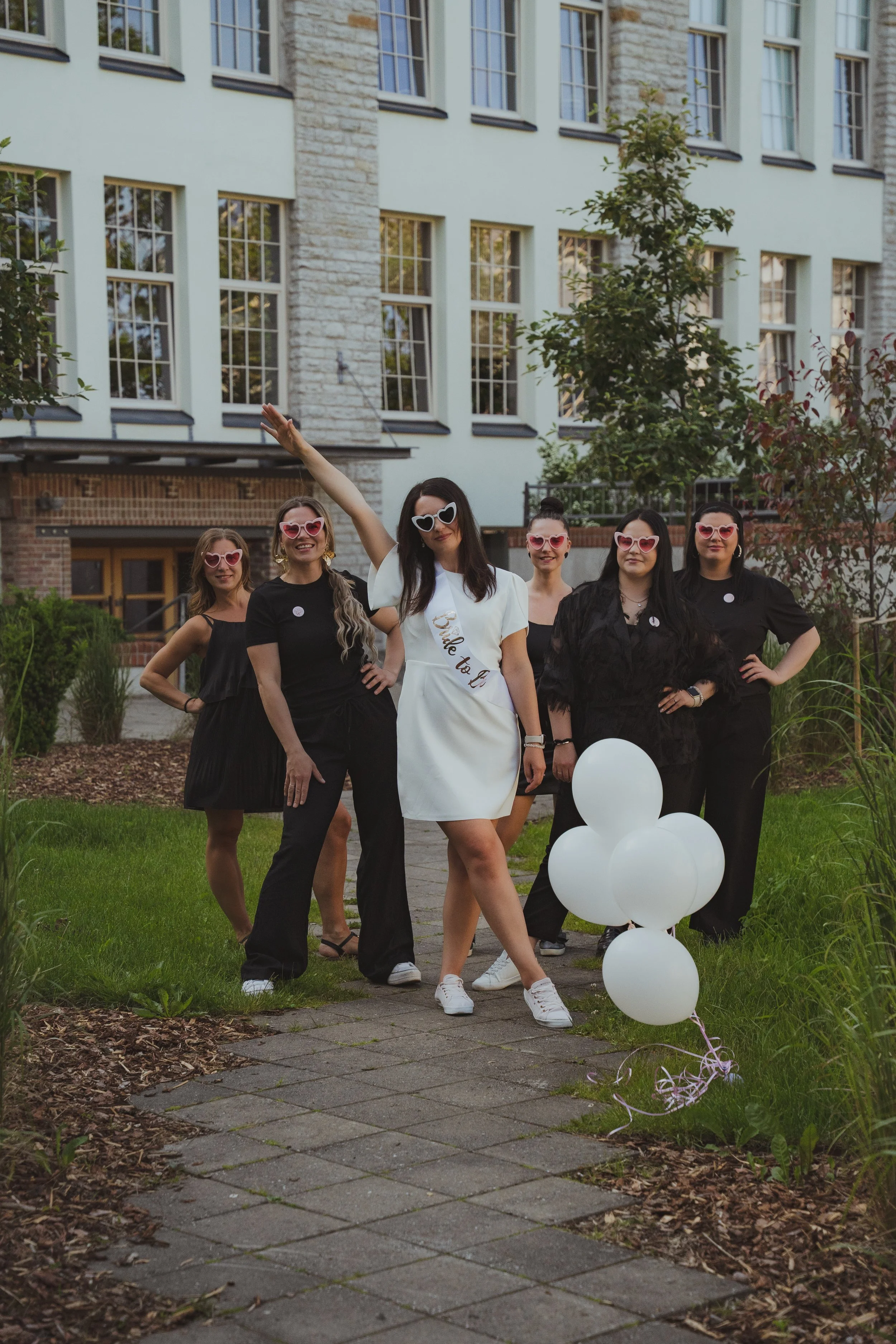 A group of women celebrating with a bride-to-be, all wearing heart-shaped sunglasses, standing outdoors on a garden path with a white building behind them. The woman in the center is wearing a white dress with a sash that reads "Bride to Be" and is holding white balloons.