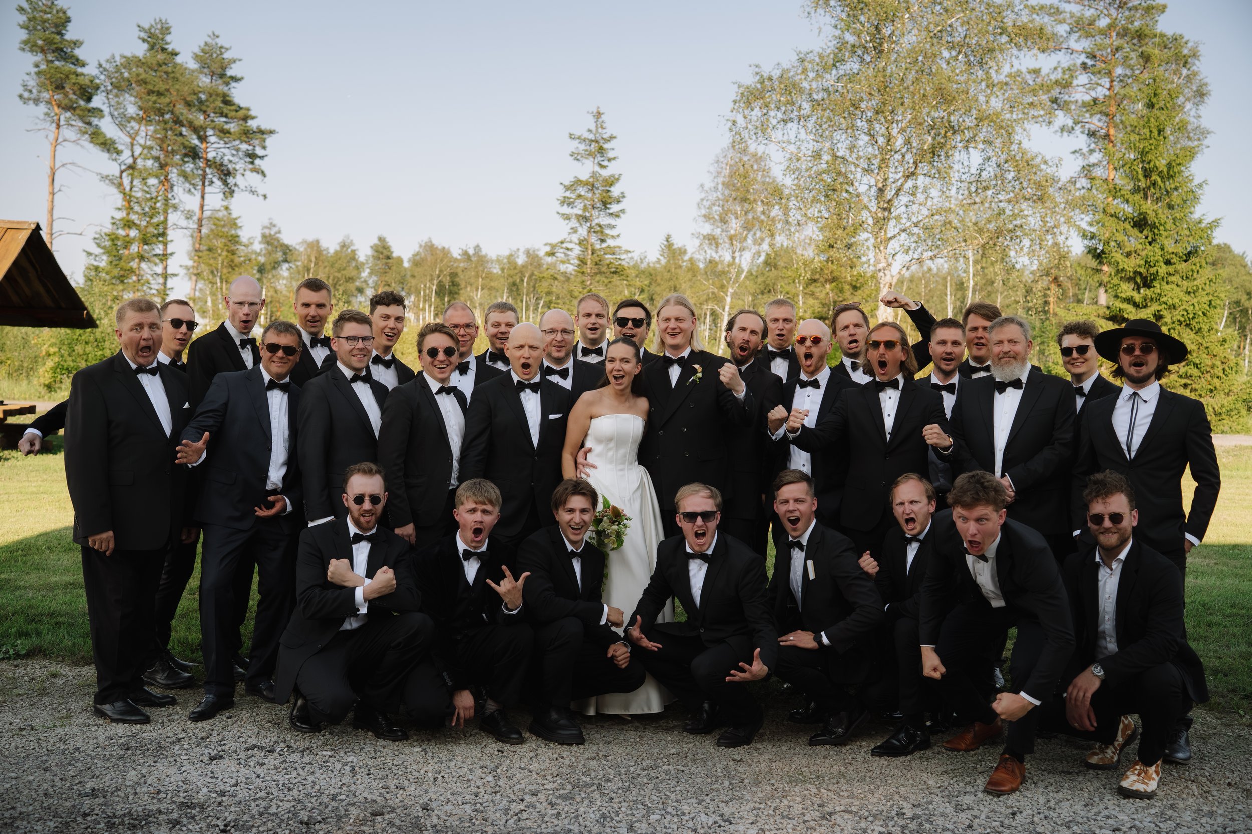 A group of men in tuxedos and a woman in a wedding dress posing outdoors, some making playful gestures and expressions, with trees and a grassy area in the background.