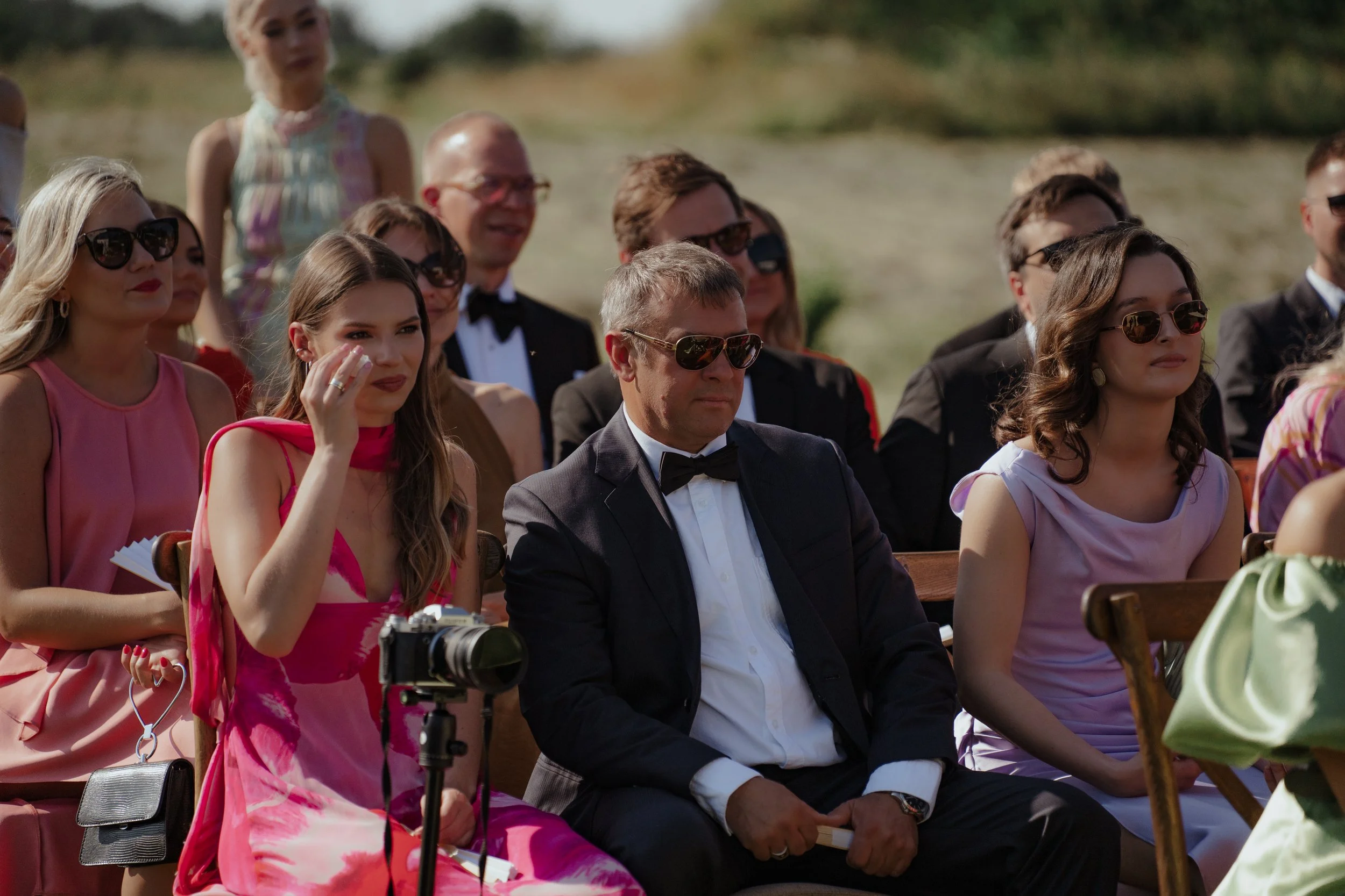 People sitting outdoors attending a formal event, with men in tuxedos and women in elegant dresses, some wearing sunglasses.