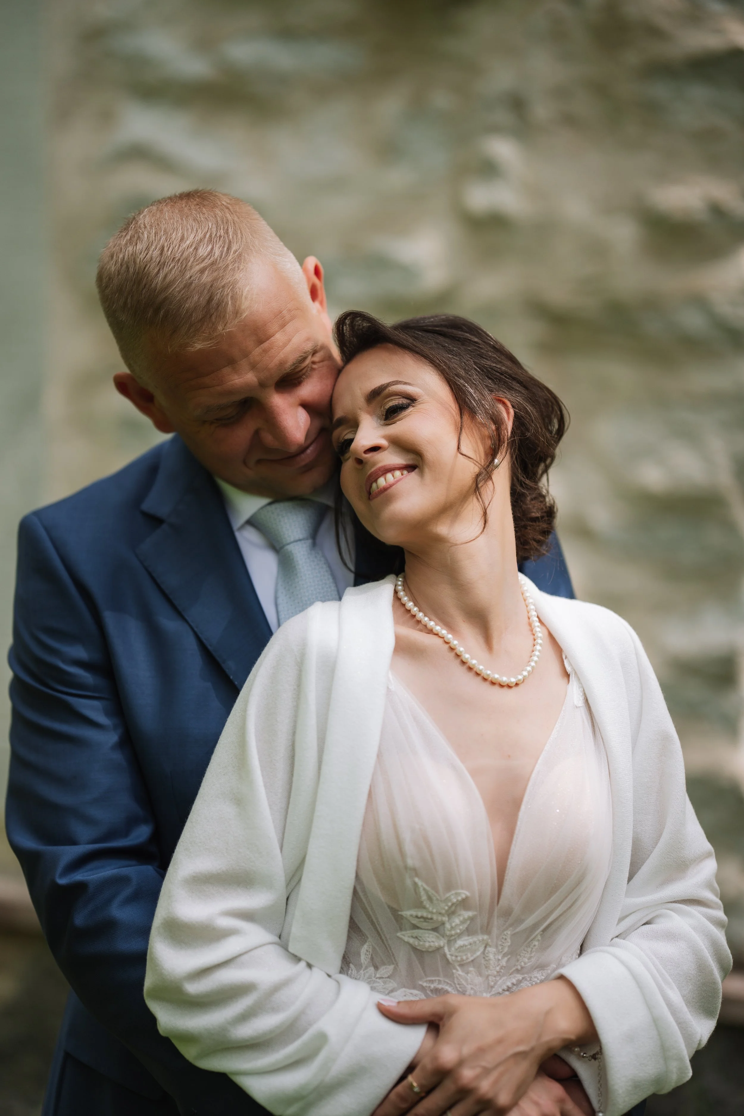 A couple dressed in wedding attire, smiling and embracing each other outdoors near a stone wall.