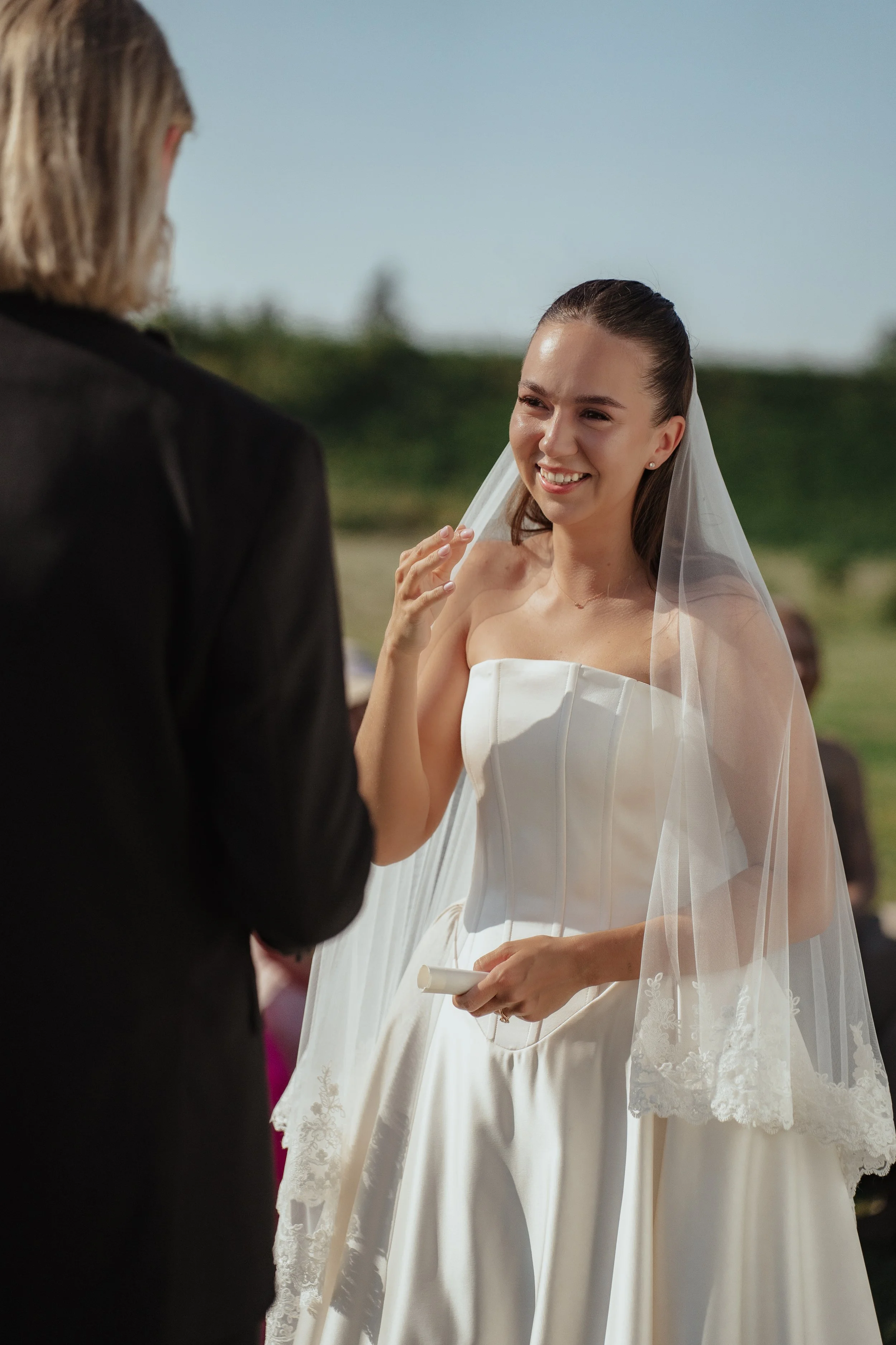 A bride in a white wedding dress with lace details on the veil, smiling as she holds a ring box, during her wedding ceremony outdoors.