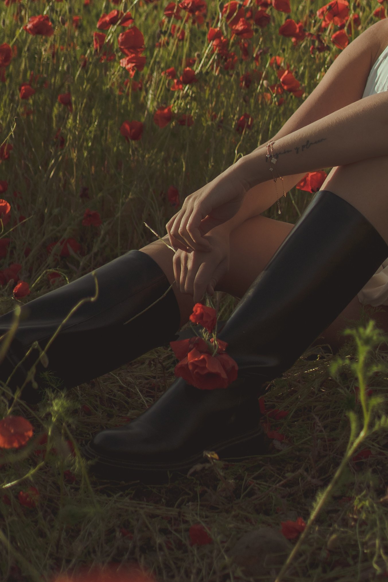 A person sitting in a field of red poppies wearing black rain boots and a white top, with arms crossed over knees and a bracelet on one wrist.