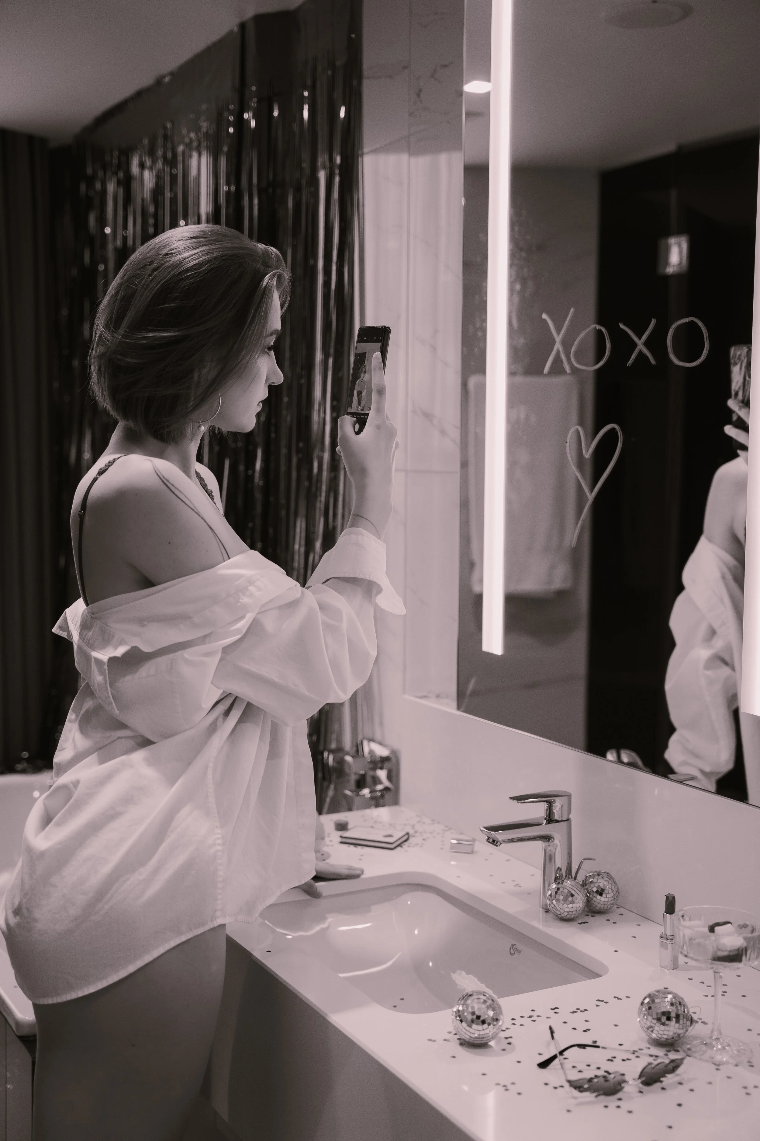 A woman standing in front of a bathroom mirror taking a selfie with her phone. She is wearing an off-shoulder shirt and has short hair. The mirror has 'XOXO' and a heart drawn on it with lipstick. The bathroom counter has disco balls, glitter, and makeup items, with a sink and faucet visible.