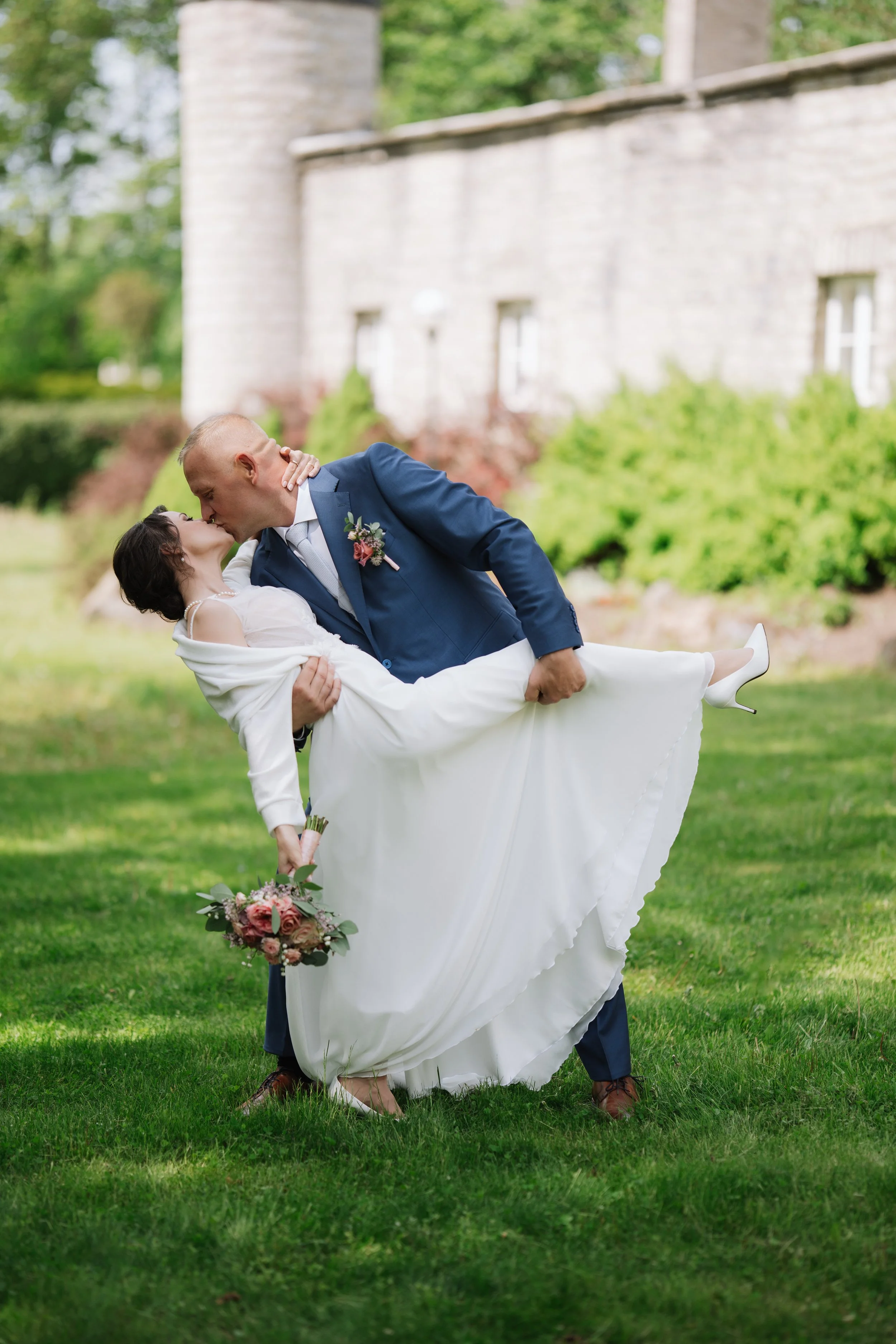 A groom in a blue suit carries a bride in a white wedding dress and heels, who is holding a bouquet of roses, as they share a kiss outdoors on a grassy lawn with a stone building and trees in the background.